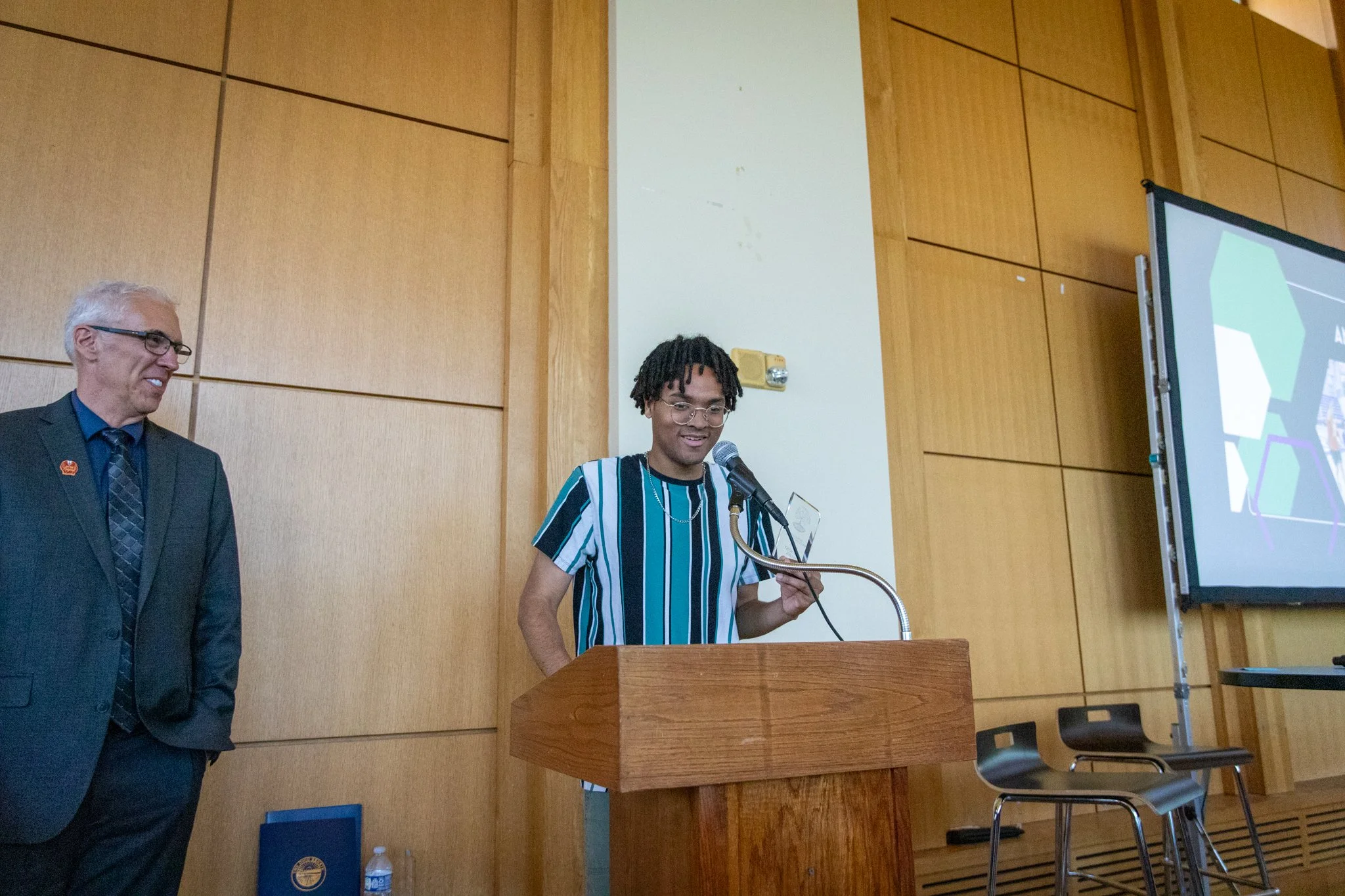 young man stands behind wooden podium speaking into a microphone