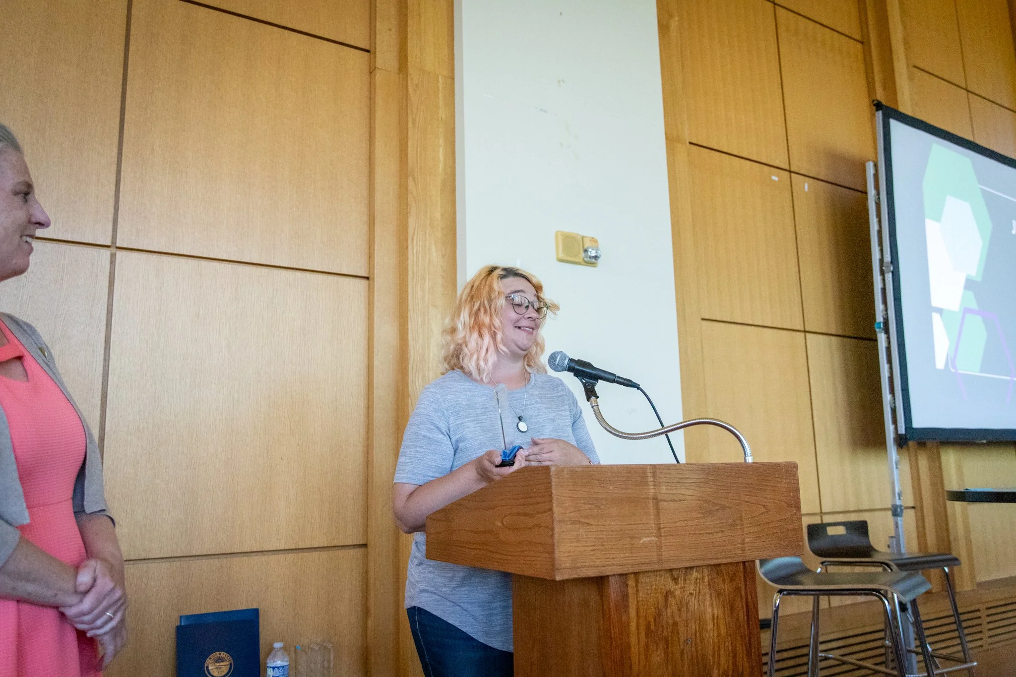 Person stands at a podium holding a small award while speaking into a microphone as another person stands nearby.