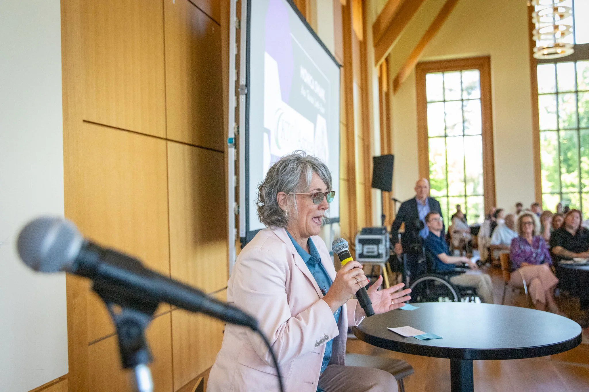 Person in a light blazer speaks into a handheld microphone at a round table while an audience listens in a bright, wood-paneled event space.