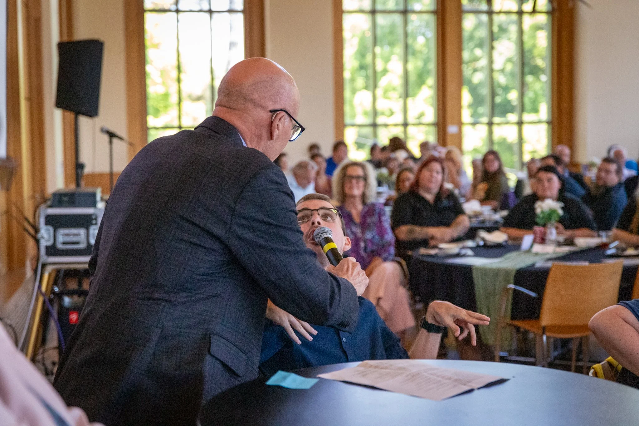 Person in a suit holds a microphone toward a seated attendee who is speaking, with an audience watching in a bright event space.