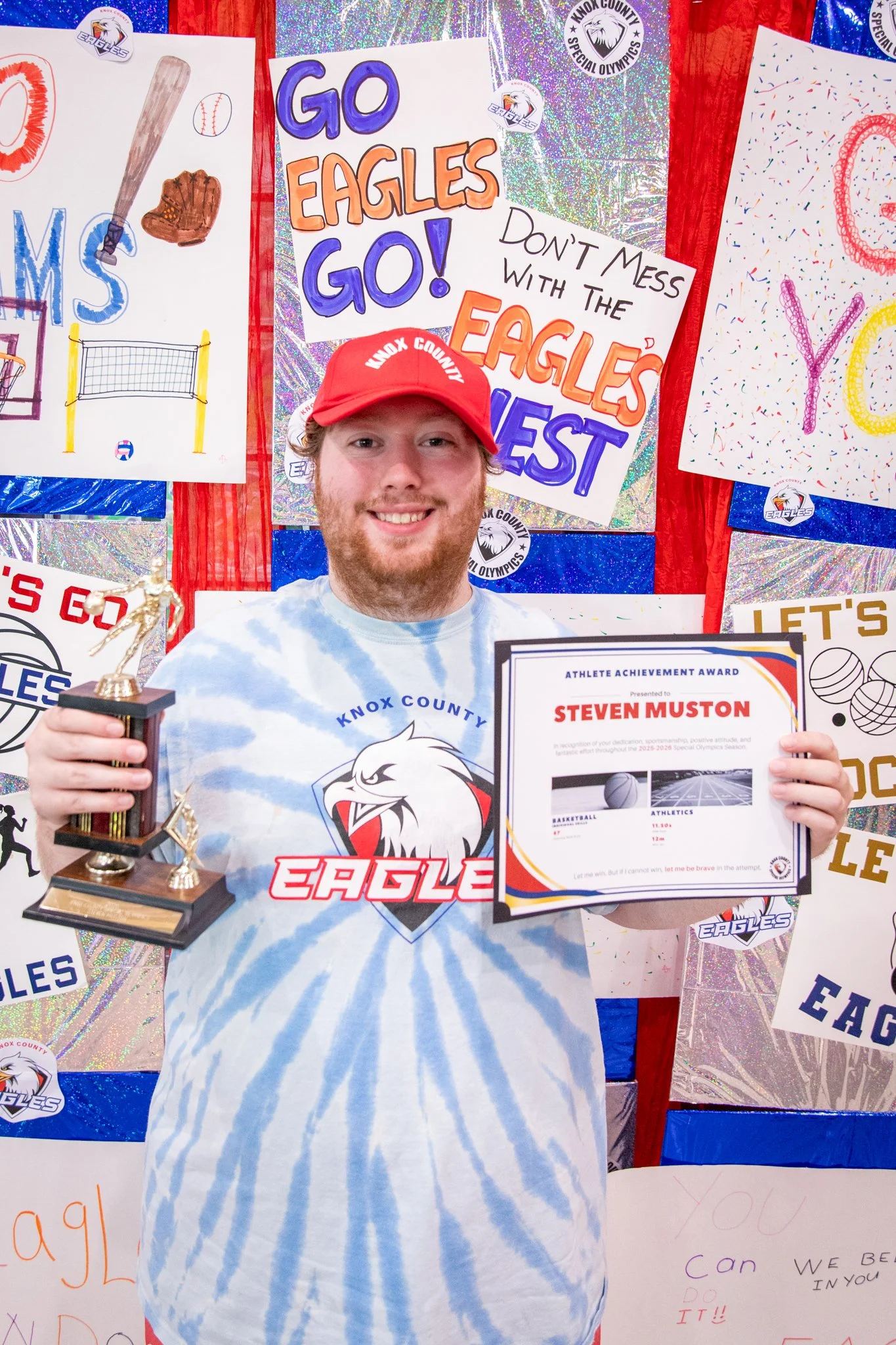 portrait of a person standing in front of a special olympics back drop, holding their trophy and certificate