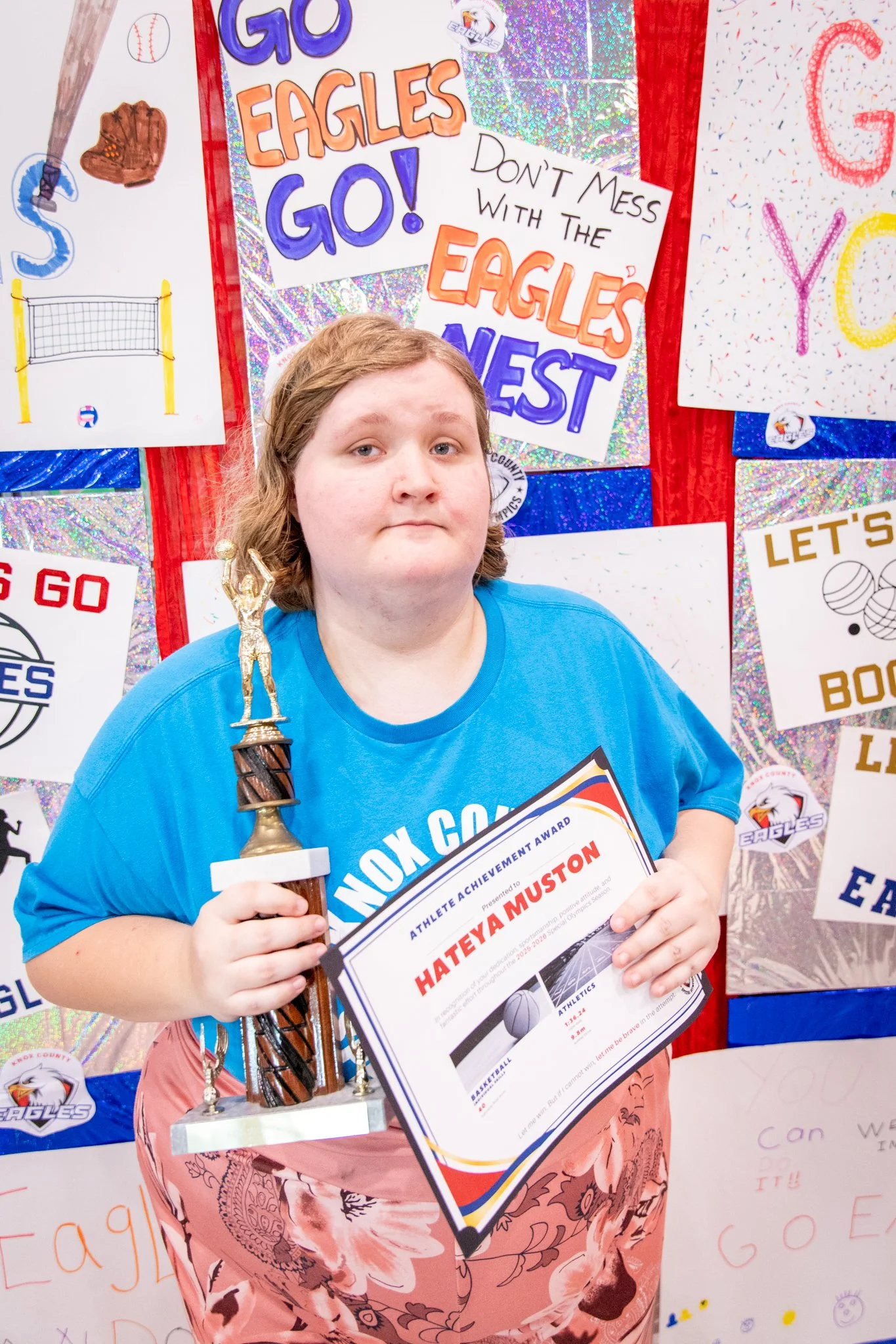 portrait of a person standing in front of a special olympics back drop, holding their trophy and certificate