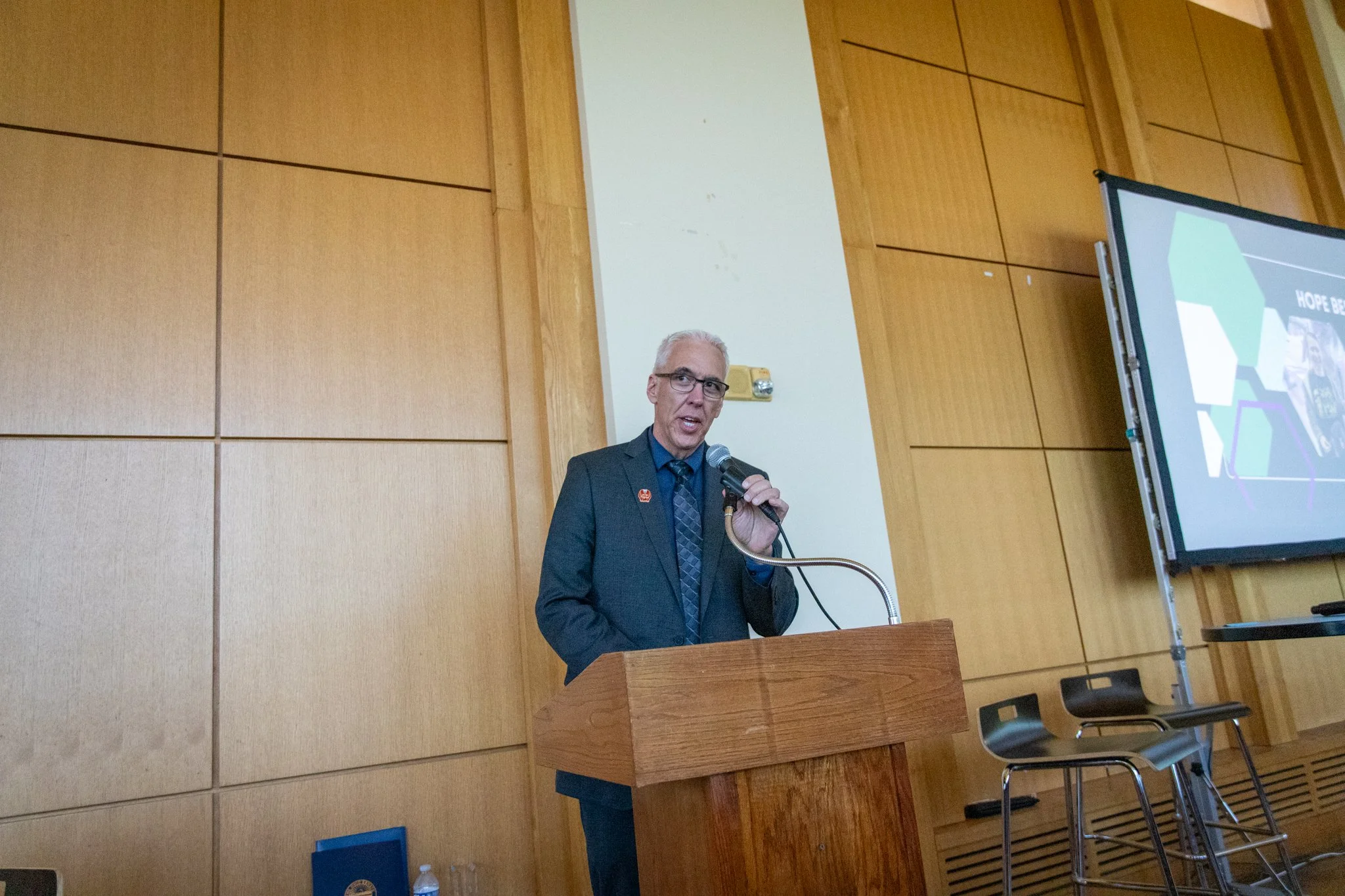Person in a suit stands at a podium speaking into a microphone in a wood-paneled event space.