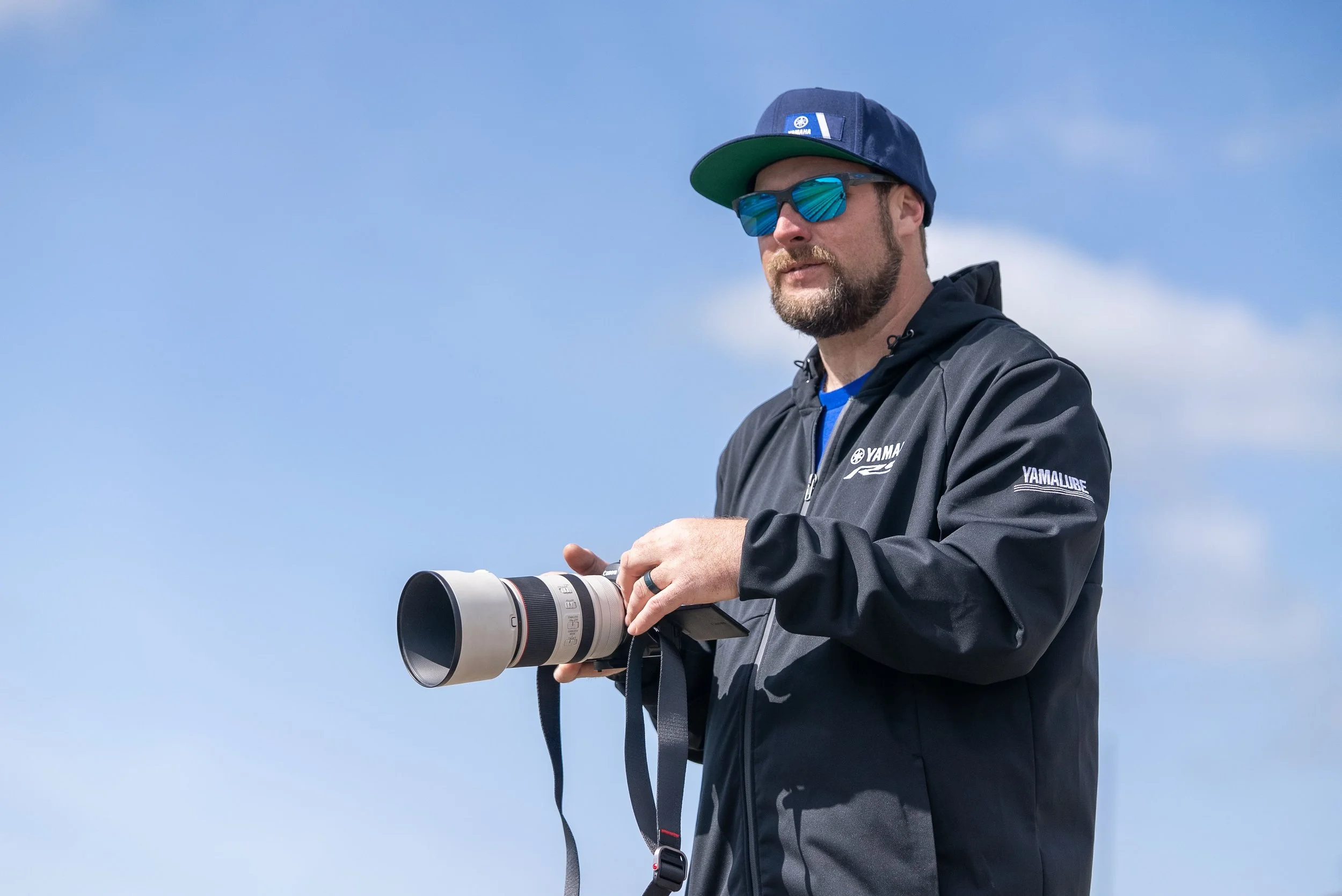 Man wearing a Yamaha jacket, blue cap, and sunglasses holding a camera with a large telephoto lens outdoors against a blue sky with clouds.