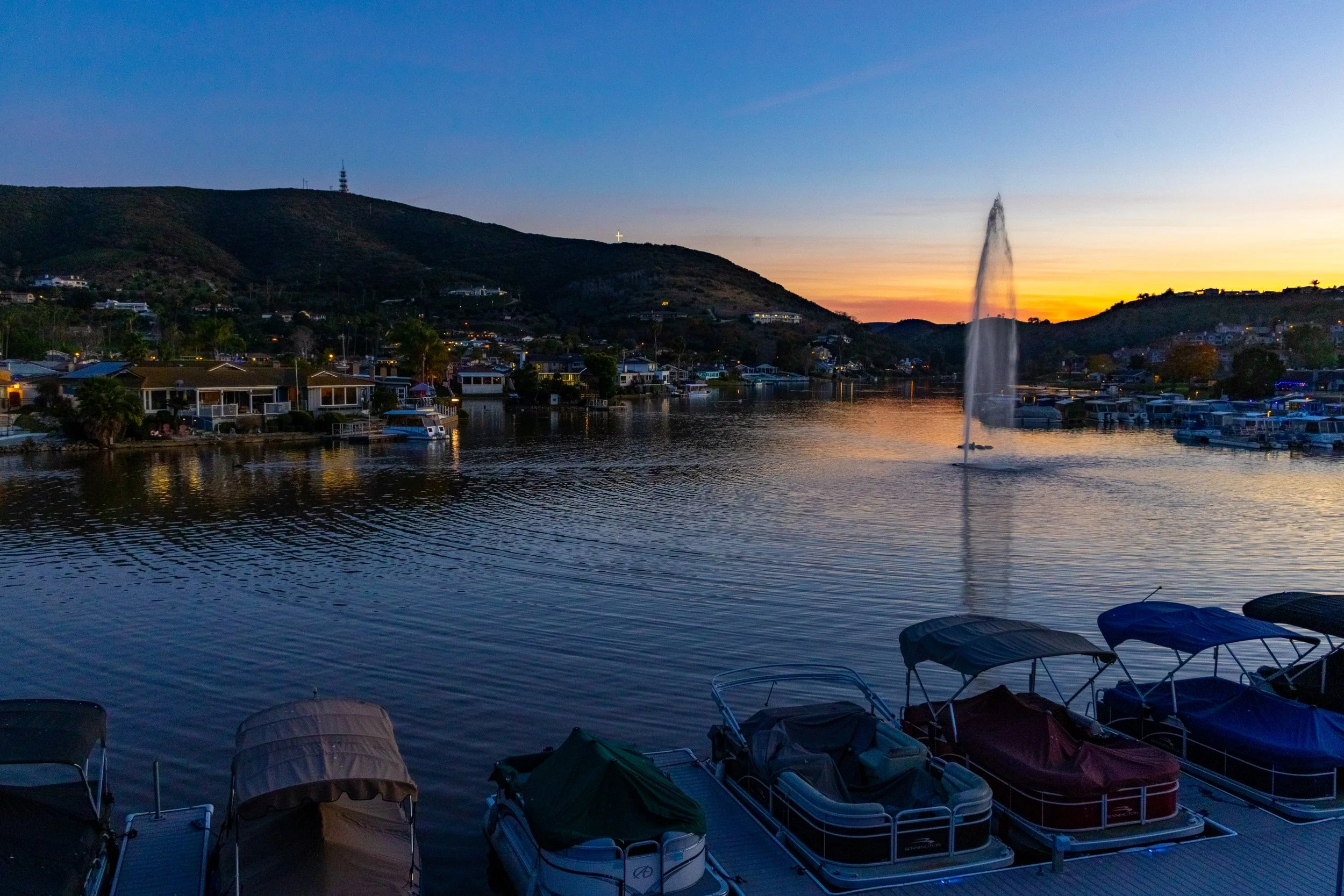 A waterfront scene at sunset with boats docked at a marina and a fountain spraying water in the lake, with residential houses and hills in the background.