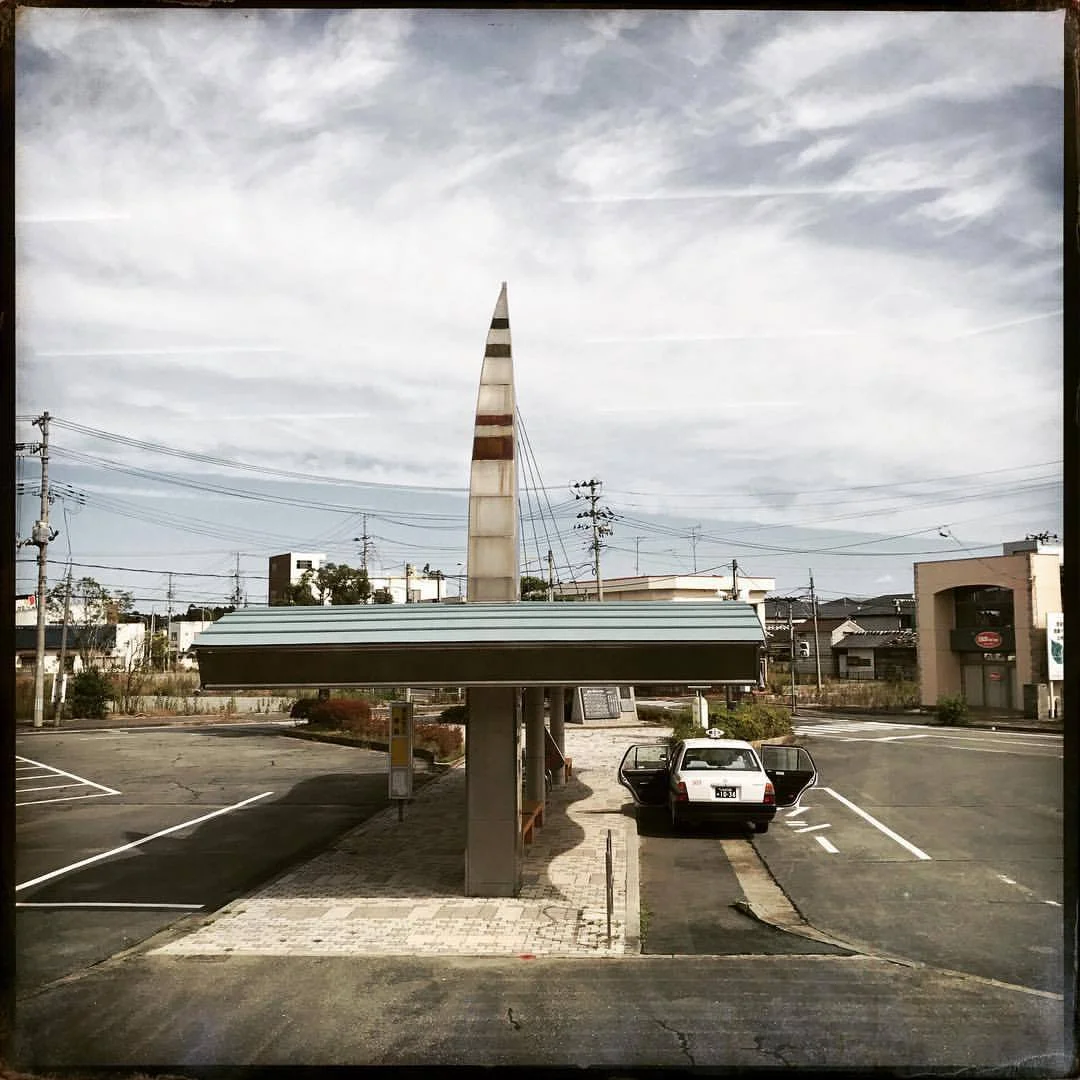 October 5, 2017 #Fukushima update: I revisited #Namie this morning, a strange experience because it’s the first time that I could go there with its new status as an open town. When it reopened in April of 2017 there was a lot of fanfare about it—but the decision to open it seemed like an act to have people forget about the disaster more than anything else. By opening it, the suggestion is that everything is back to normal and therefor people should not worry any longer about what happened. Open status aside, it really is still a closed off place because in the time it was within the exclusion zone buildings crumbled and people who owned businesses moved—only 2% of the population have returned. Many of the elderly who might have returned sadly did not make it long enough to see their town “reopen.” …And on top of it, higher than normal amounts of radiation persist. Namie feels lonelier open than closed. When it was off limits and a part of the exclusion zone it existed as a representation of a place that once was more than actually being a place itself. The town represented a sad lament, it represented something that was once connected to life. In that time of lament there were still the old crumbling buildings with their hand painted and fading signs—I could feel the past in these withering structures. I could hear the conversations and clatter of classes that filled the bars and izakayas. Many of those establishments are gone now. Torn down, the space where they used to be are now austere flattened lots, antiseptic places that reveal nothing about the past and offer nothing for the future. Of the 18,000+ residents who used to live in Namie about 280 have returned. I walked past all of the main streets and did not see one open business aside from the post office, some administrative places and the gas station that is mostly there to service work vehicles. There’s a little food court along the main highway, but it did not look very appealing. Apart from those businesses, from what I understand, there are no social services or even places to get decent food nearby. Just outside the station, a lone taxi driver waited for a passenger to come off from one of the semi hourly trains, doors open as you’d find in many small towns here. But the train arrived empty and it left empty. The taxi driver, with nowhere to go, kept idling outside of the station reading his newspaper and waiting for customers in a town largely without people. — at Namie Station .