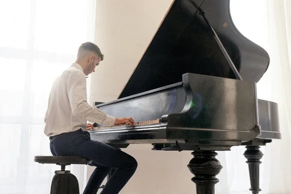 A man in a white shirt and dark pants playing a grand piano in a room with large windows.