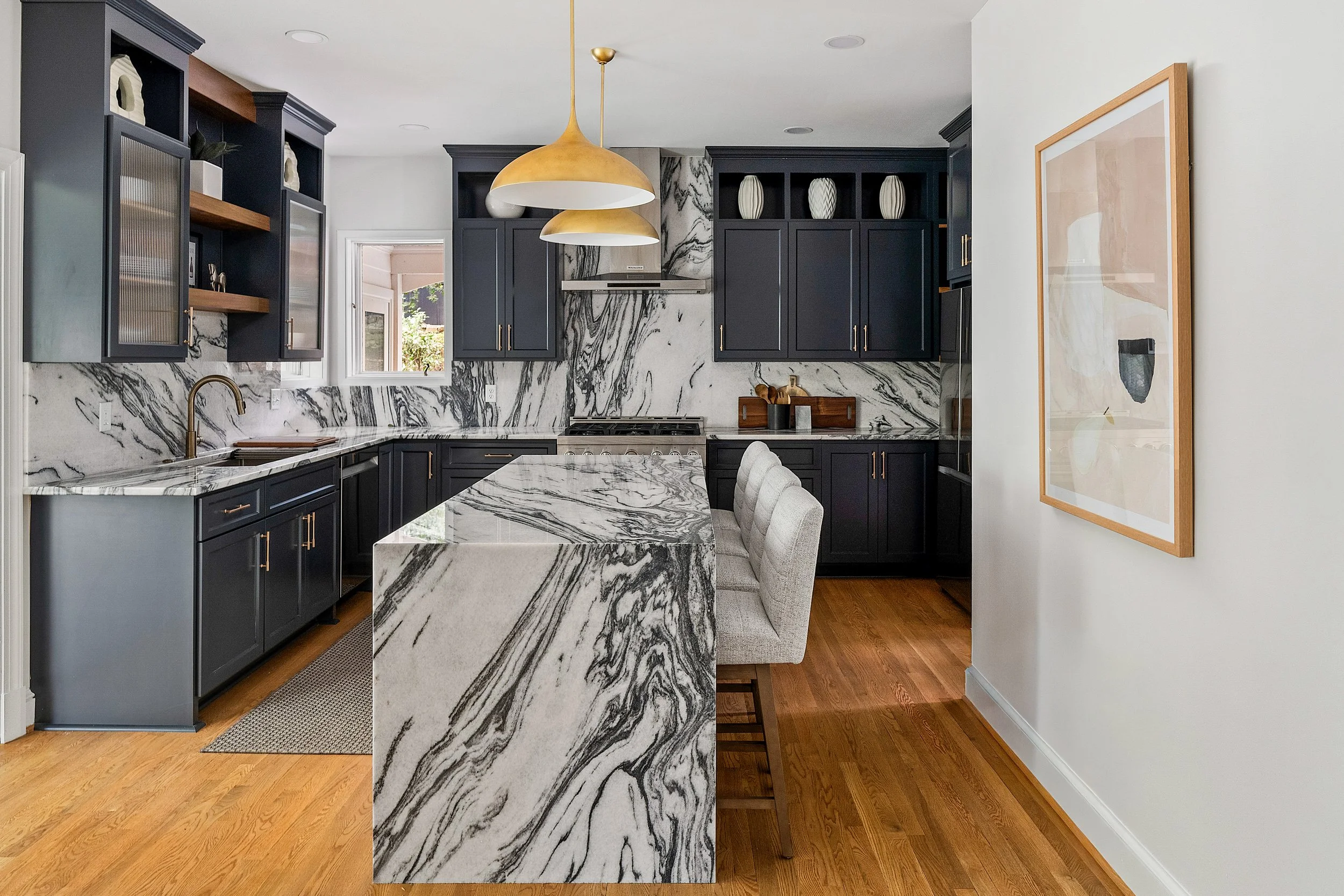 Modern kitchen with dramatic blue cabinetry, waterfall marble island, brass pendant lighting, and full-height marble backsplash in a luxury home.