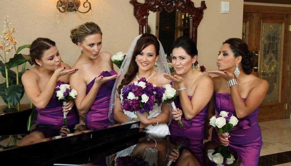 A bride in a white wedding dress holding a bouquet of purple and white flowers, surrounded by four bridesmaids in purple dresses, each holding a similar bouquet, in a decorated indoor setting.