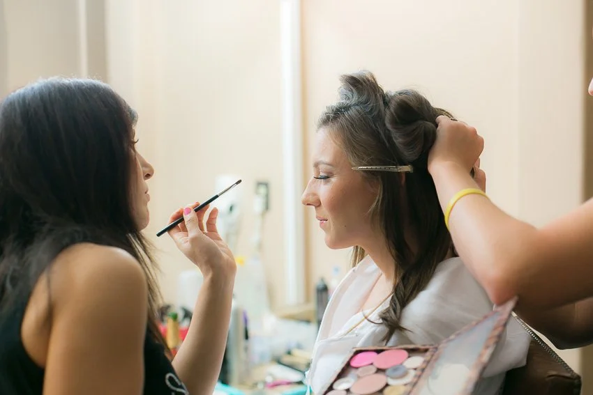 A woman getting her makeup done, with her eyes closed, while a makeup artist applies makeup and styles her hair.