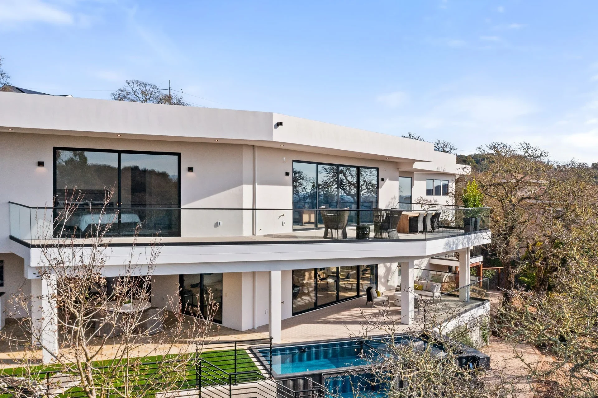 Modern multi-level contemporary custom house with large windows and balconies, outdoor patio with seating, and a swimming pool, surrounded by trees under a clear blue sky.