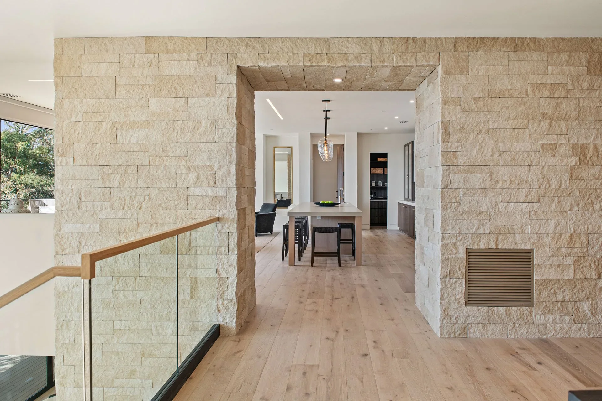 Open-concept kitchen and living space with beige stone walls, light-colored hardwood floors, a central island with barstools, black cabinetry, minimal decor, and large windows allowing natural light.