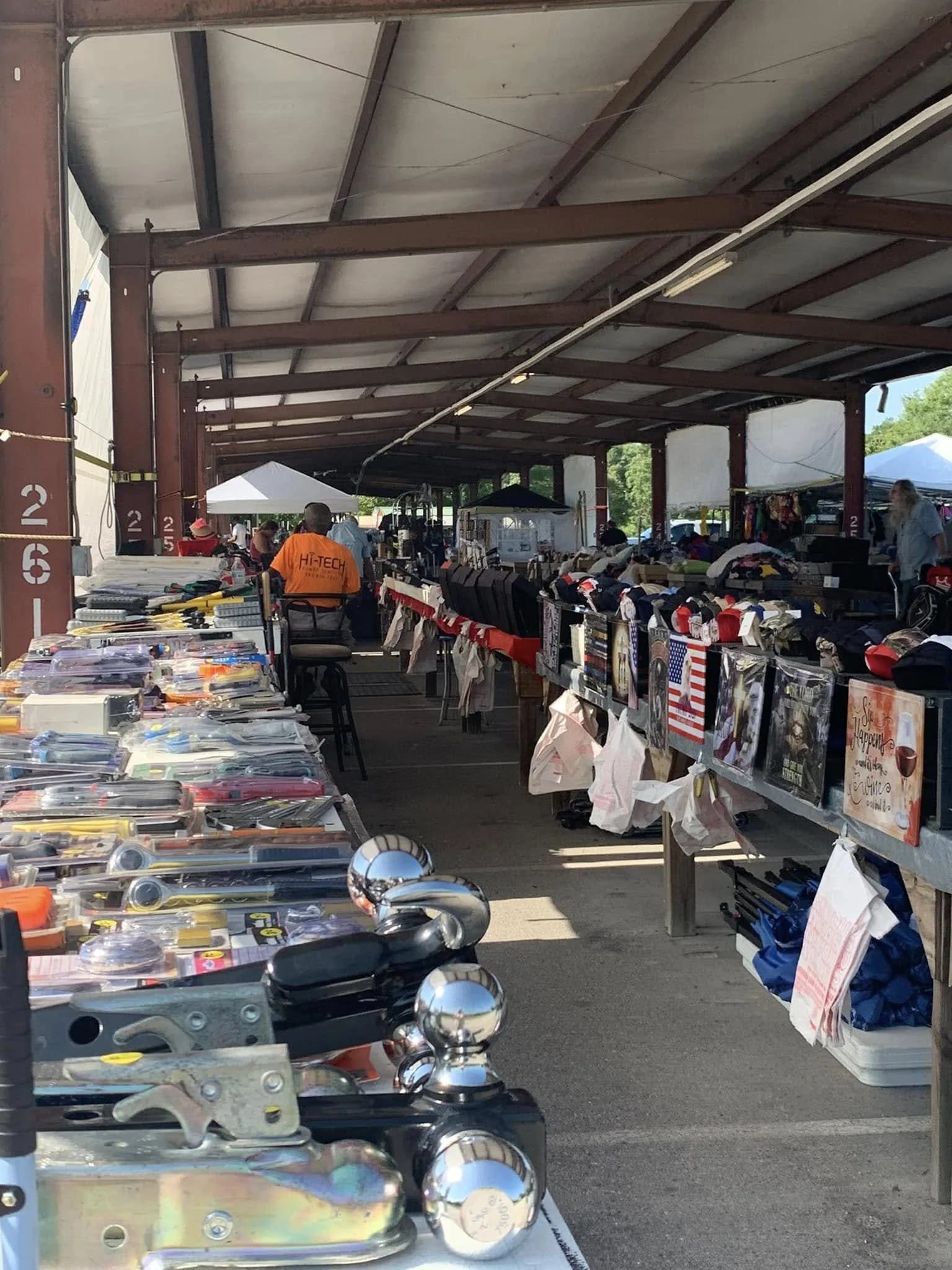 Outdoor market stall with various tools, accessories, and merchandise on tables and racks under a metal roof. People browsing and shopping.