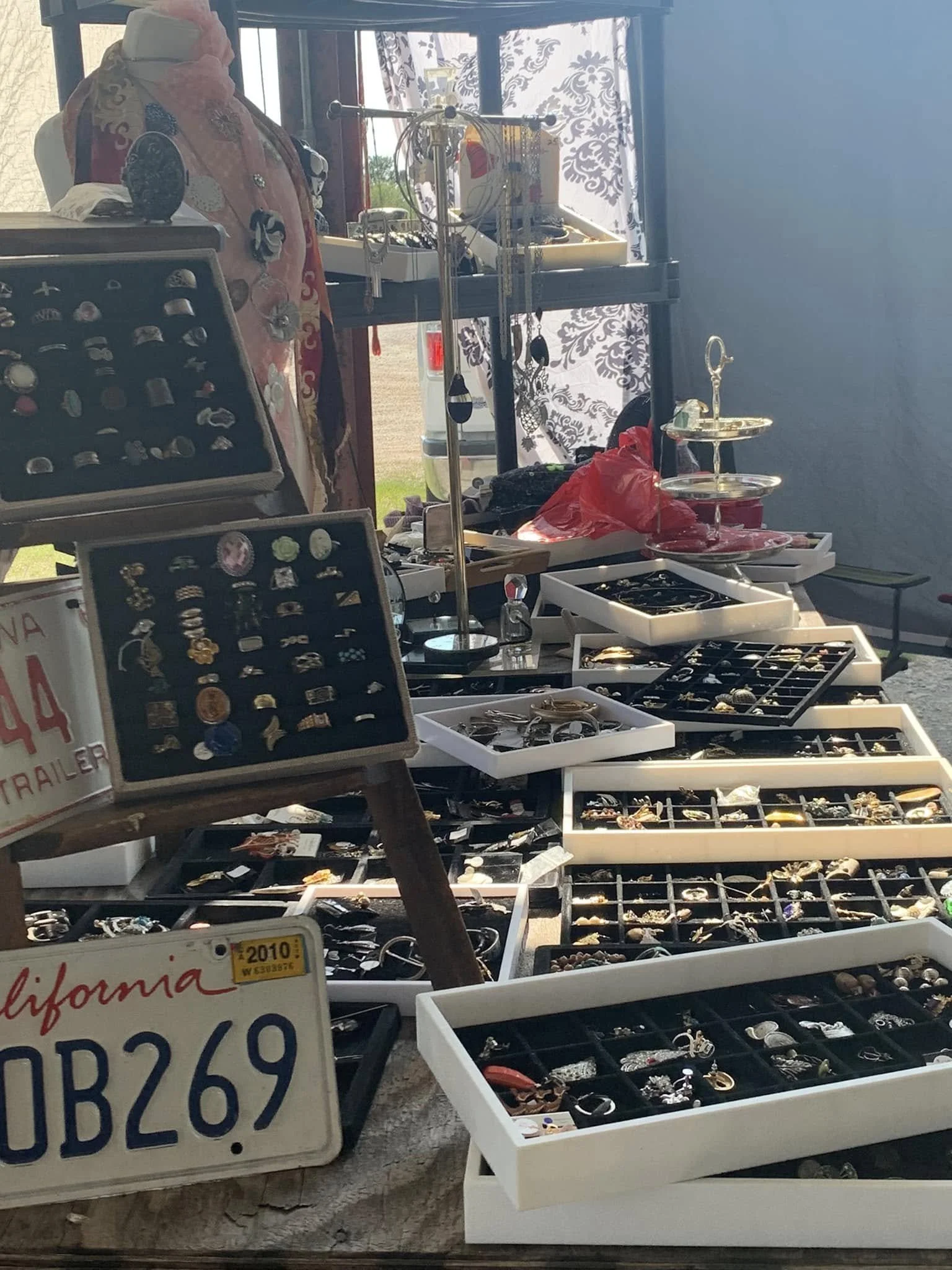 A display of jewelry and accessories at an outdoor market stand, with trays filled with rings, earrings, and bracelets, and a vintage California license plate used as decoration.