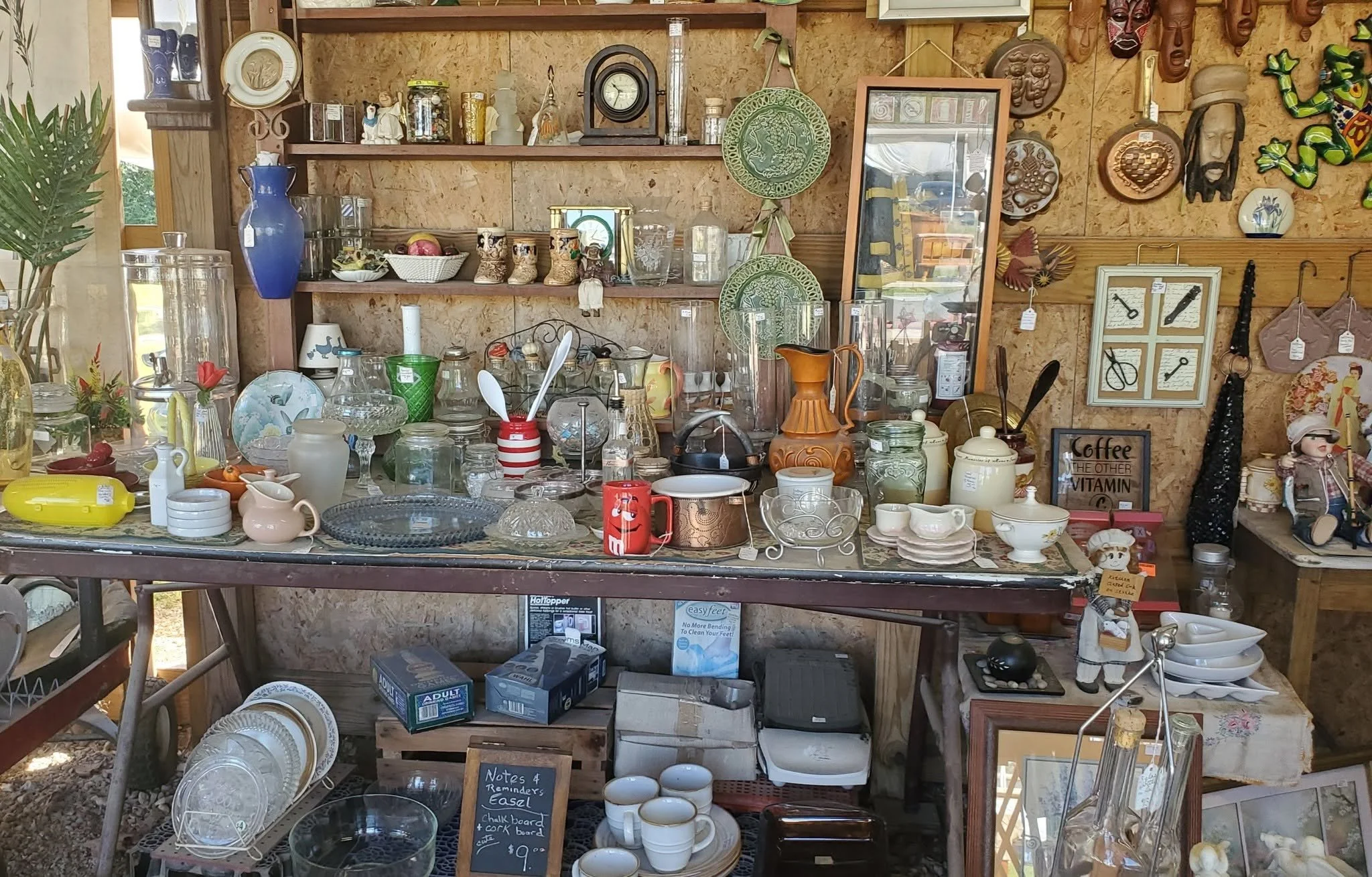 A table at a thrift store with various glassware, ceramics, and decorative items, with shelves and wall decorations behind it.