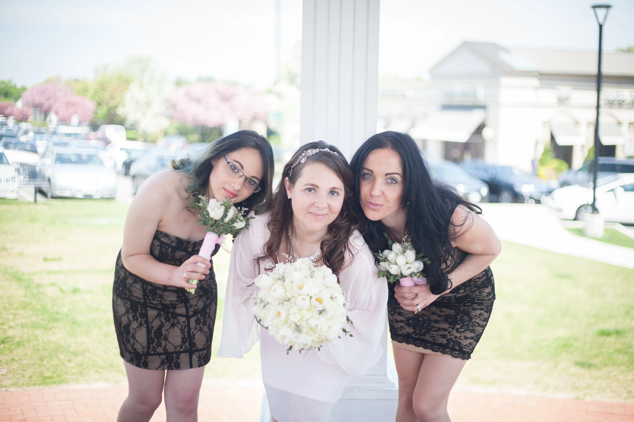 Three women, one in the middle holding a bouquet of white flowers, showing affection and friendship, outside near a parking lot with trees in bloom in the background.