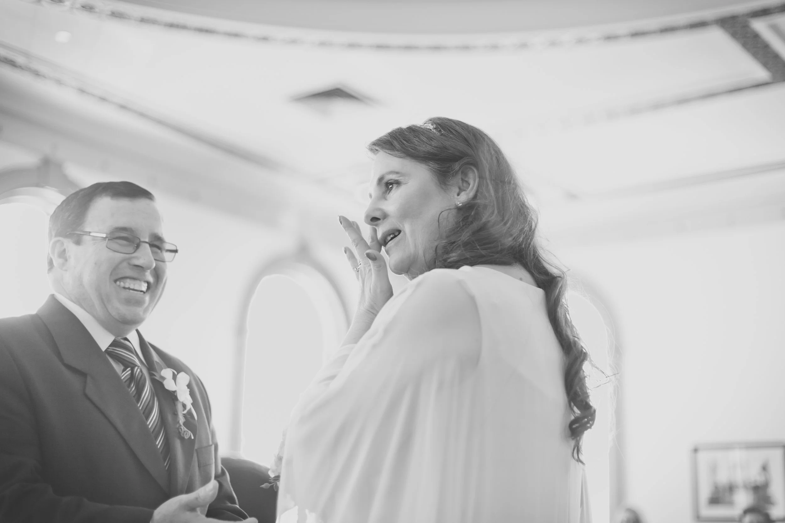 A black and white photo of a man and woman at what appears to be a formal event or wedding. The man is dressed in a tuxedo with a boutonniere, smiling and looking at the woman. The woman, wearing a dress and a headband, is gesturing with her hand nea