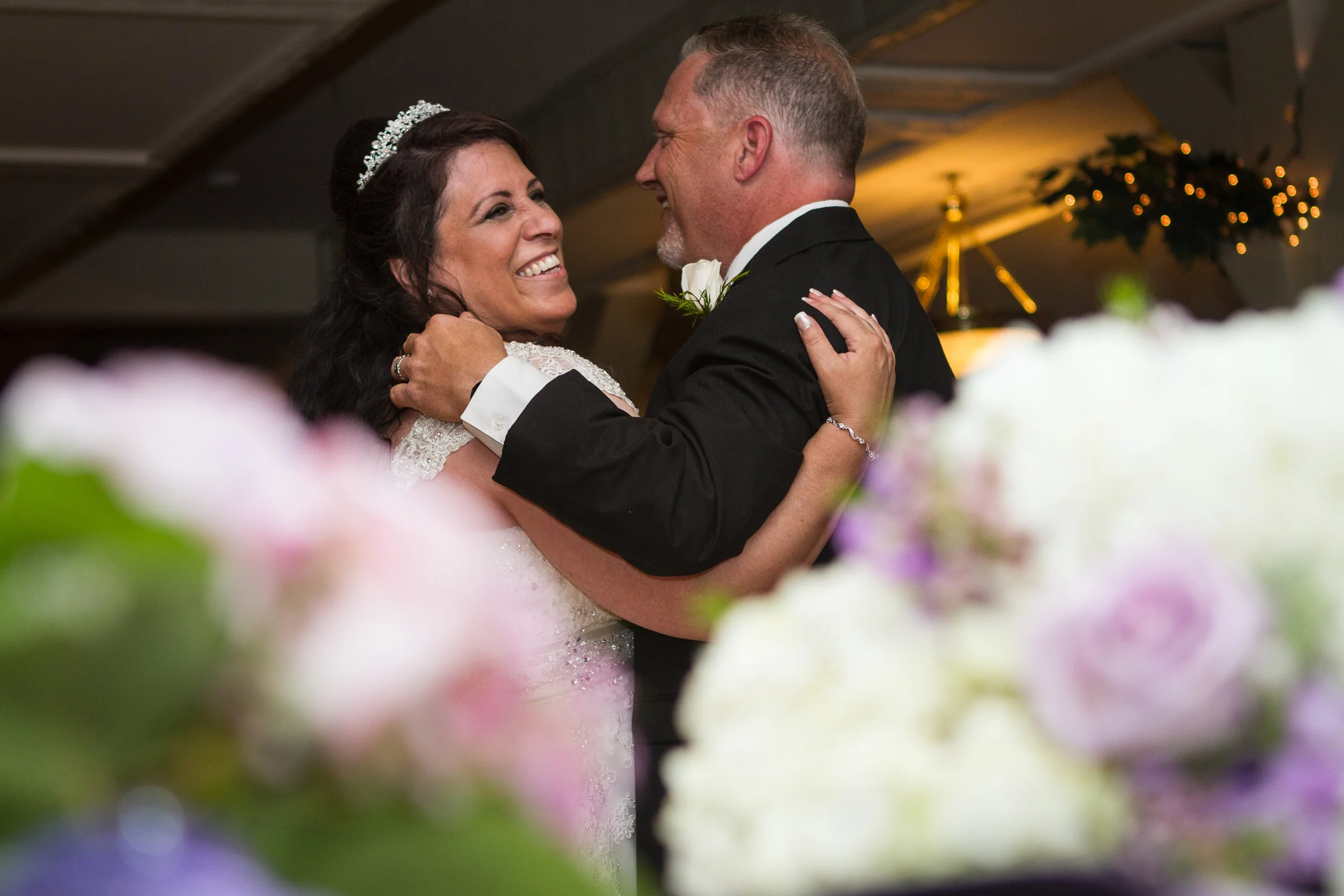 A bride and groom dancing at their wedding, smiling and looking at each other, with wedding decorations and flowers in the background.