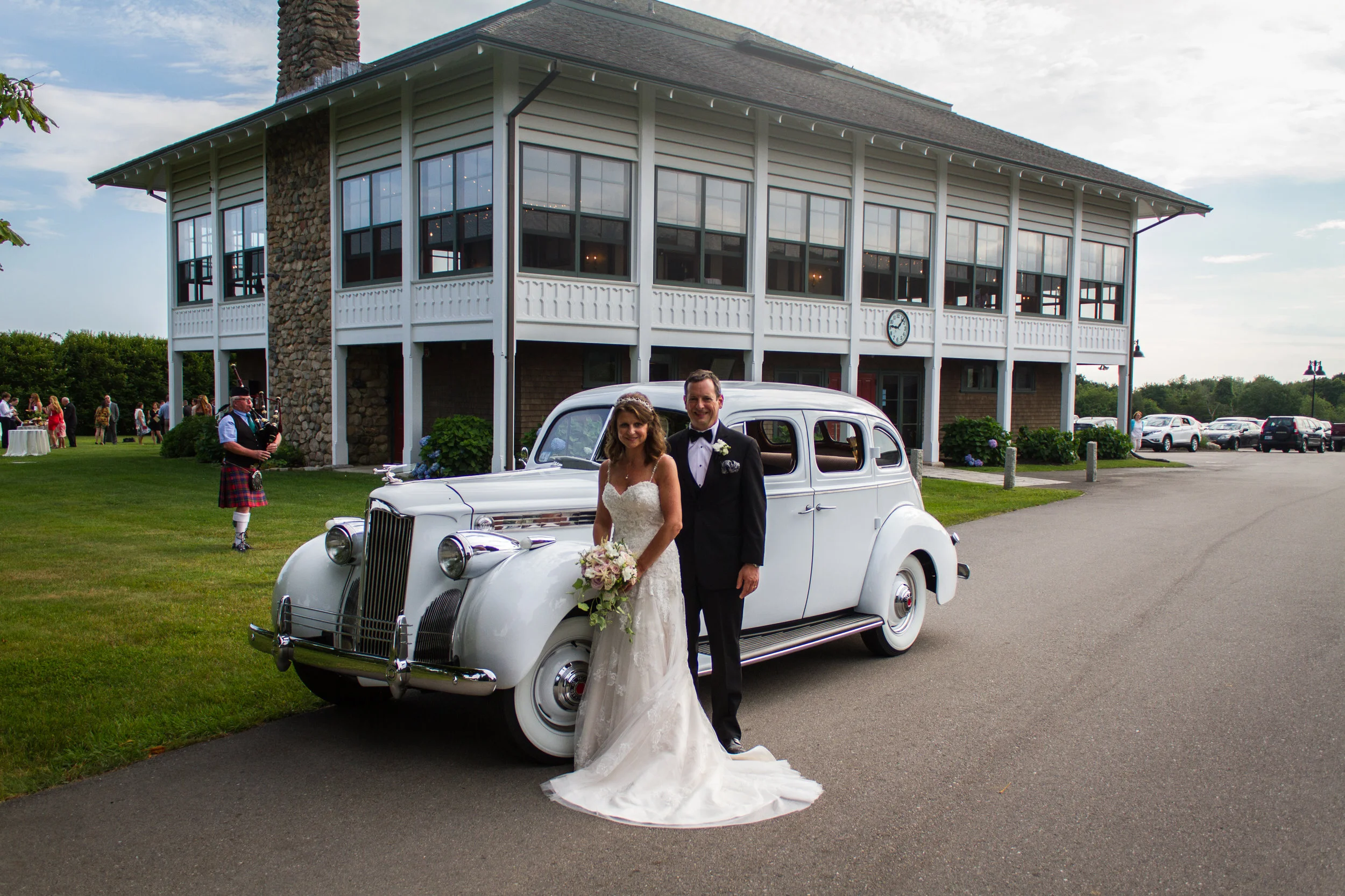 A bride and groom standing next to a vintage white car on a grassy area outside a large building. The bride is in a white wedding dress holding a bouquet, and the groom is in a black tuxedo. There are guests and musicians, including a bagpiper, in th