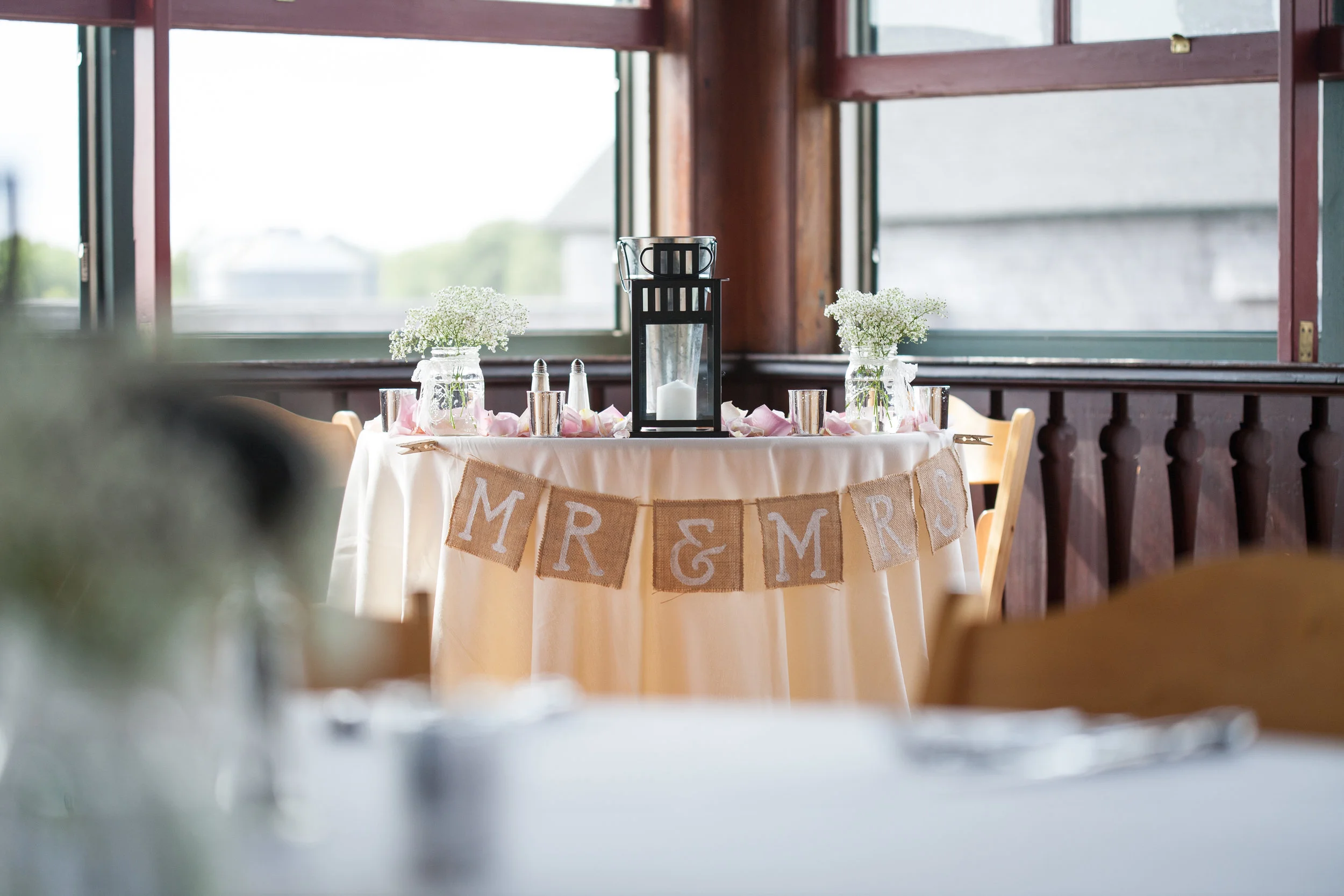 A decorated table with a white tablecloth, flower vases, candles, and a "Mr & Mrs" banner, set near large windows in a wooden room, likely for a wedding celebration.
