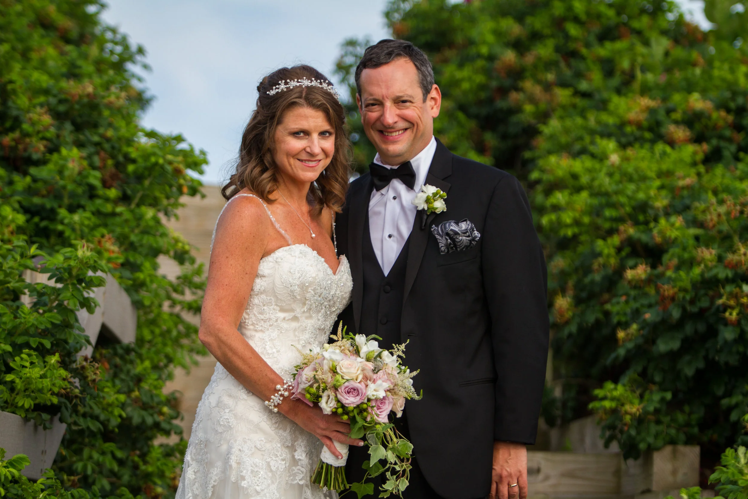 A bride and groom standing outside on their wedding day, both smiling and dressed in formal wedding attire, with the bride holding a bouquet of flowers and greenery.