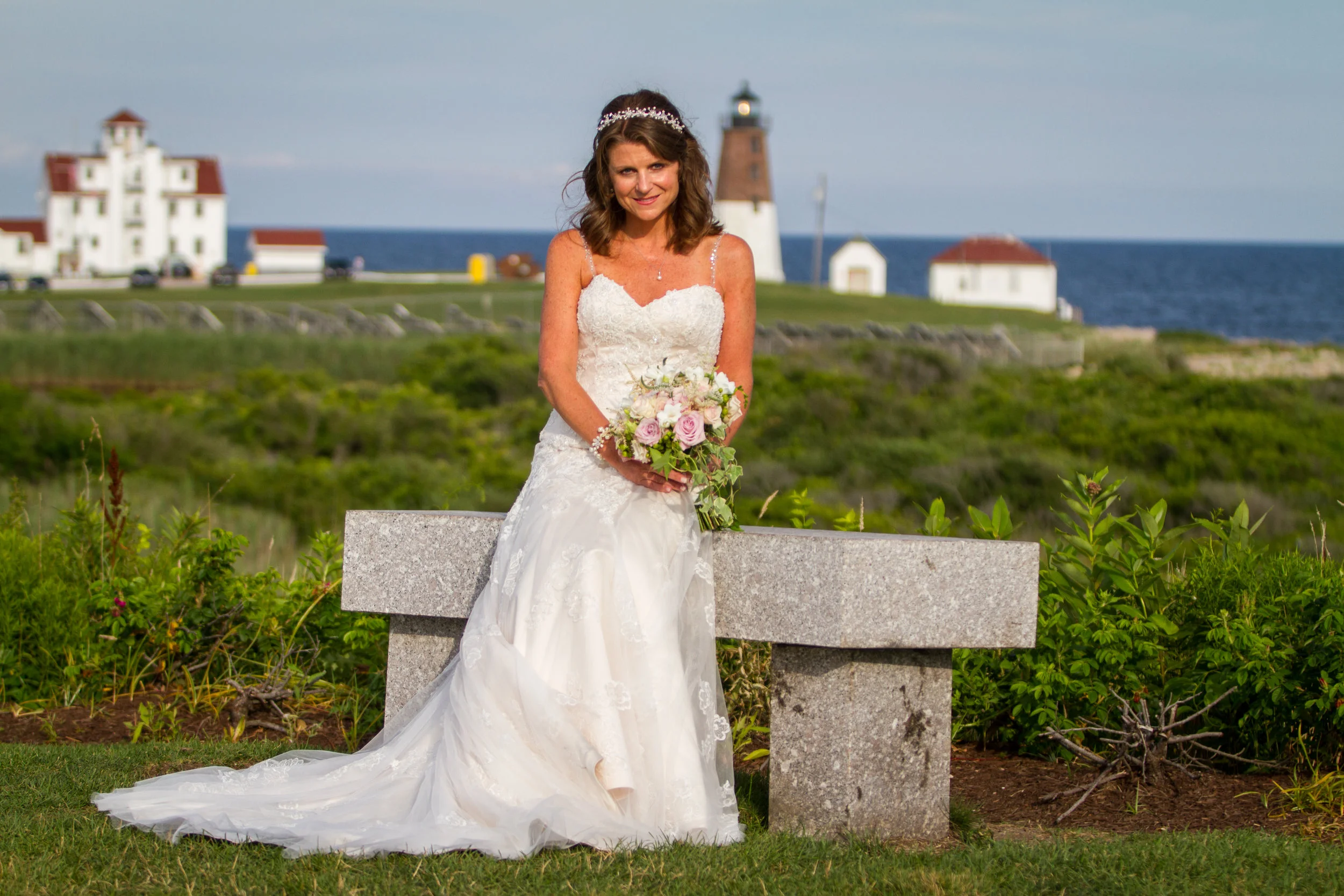 A woman in a white wedding dress holding a bouquet of pink and white flowers, standing outdoors near a stone bench with a lighthouse and buildings in the background.