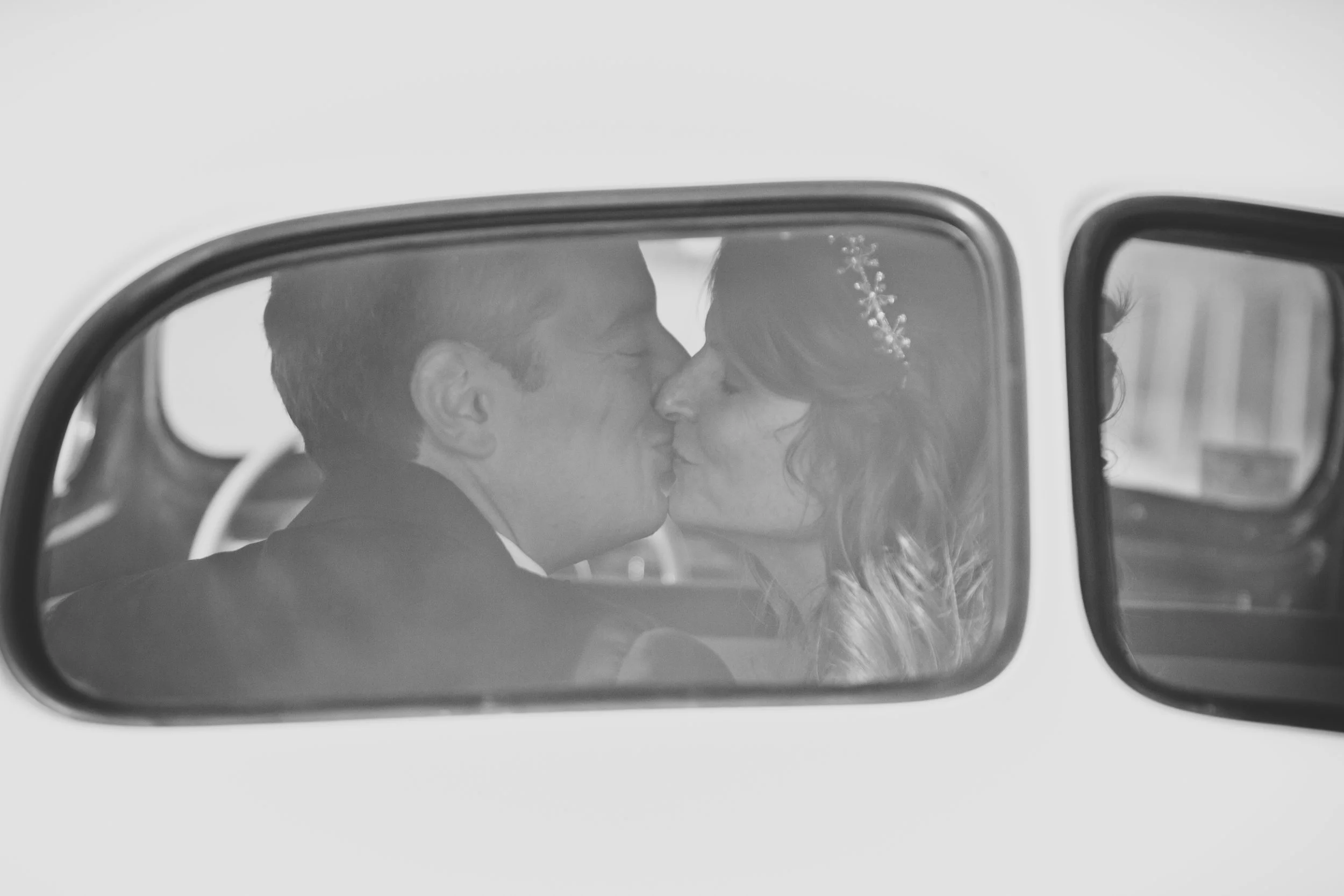 A black and white photo of a person and a woman sharing a kiss, seen through a car mirror.
