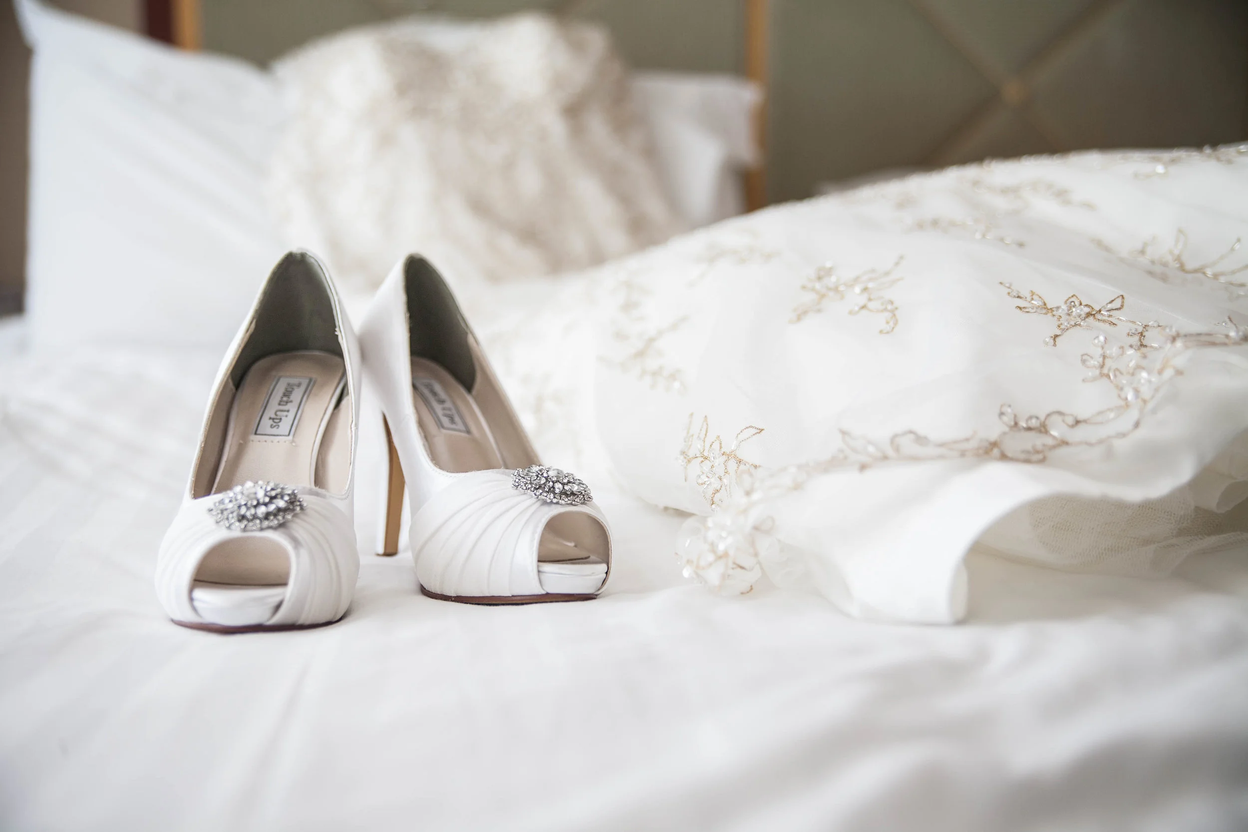 White wedding shoes with embellishments on the toes, placed on a white fabric with gold embroidery, with a pillow in the background.