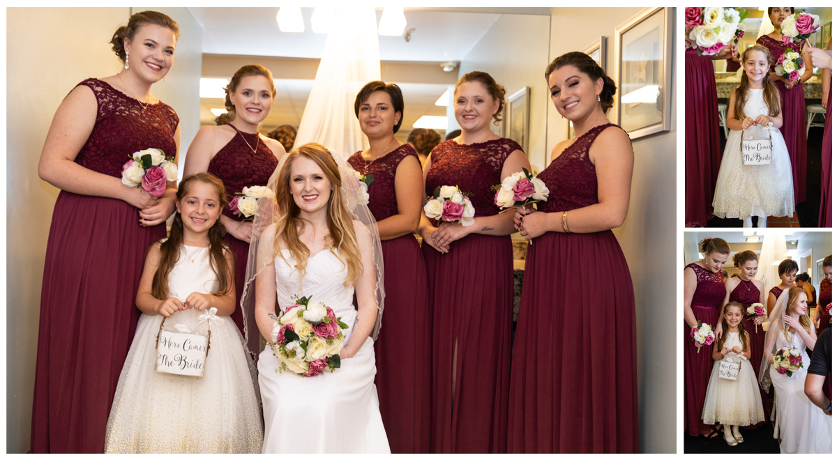 A bride with long blonde hair in a white dress holding a bouquet, five bridesmaids in burgundy dresses, and a young girl in a white dress holding a sign saying "Here Comes the Bride" at a wedding celebration.