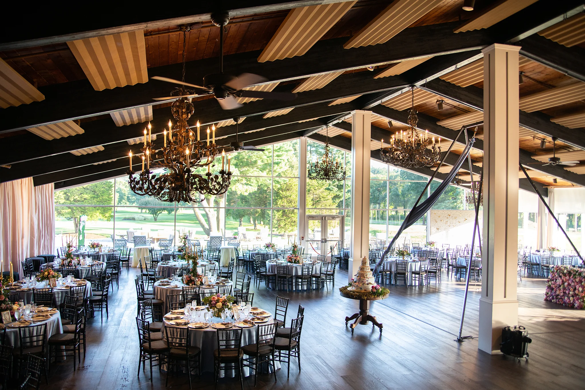 Interior of a spacious event hall with large windows, decorated for a celebration with round tables, flowers, and elegant chandeliers hanging from a wooden ceiling.