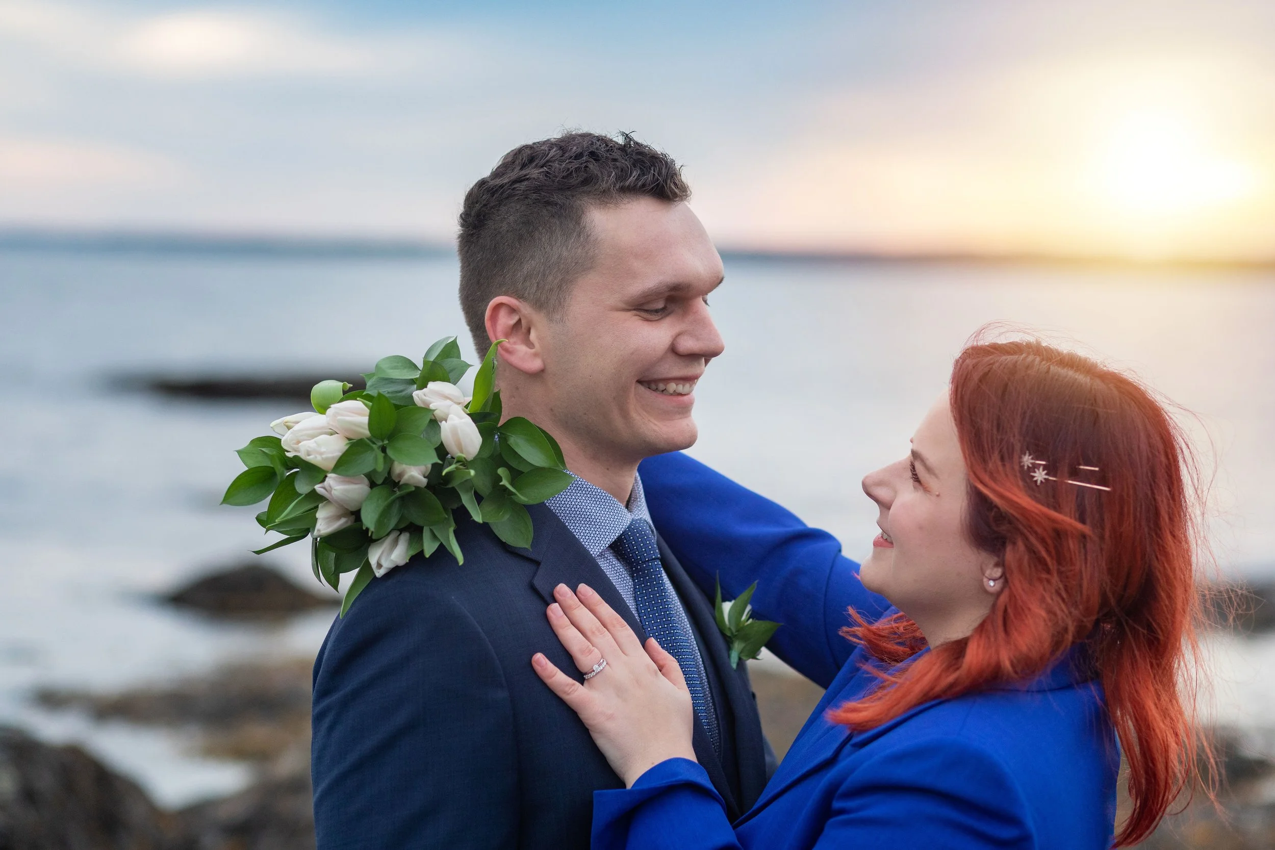 A smiling man and woman embrace outdoors near water at sunset, with the man holding a bouquet of white flowers with green leaves.