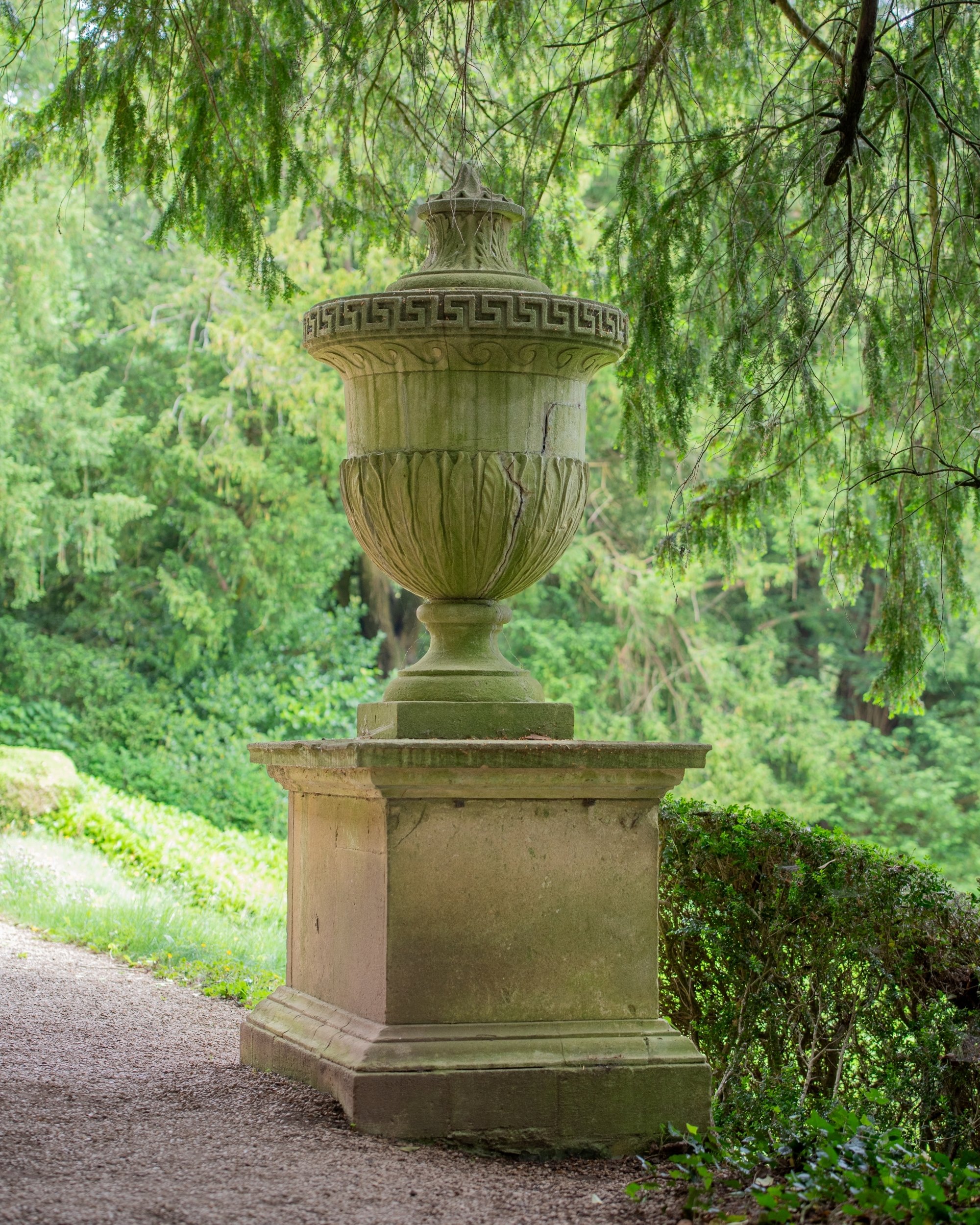 Limestone urn with a Greek key rim and carved leaf decoration on a square pedestal beside a gravel path at Rousham, framed by yew branches.