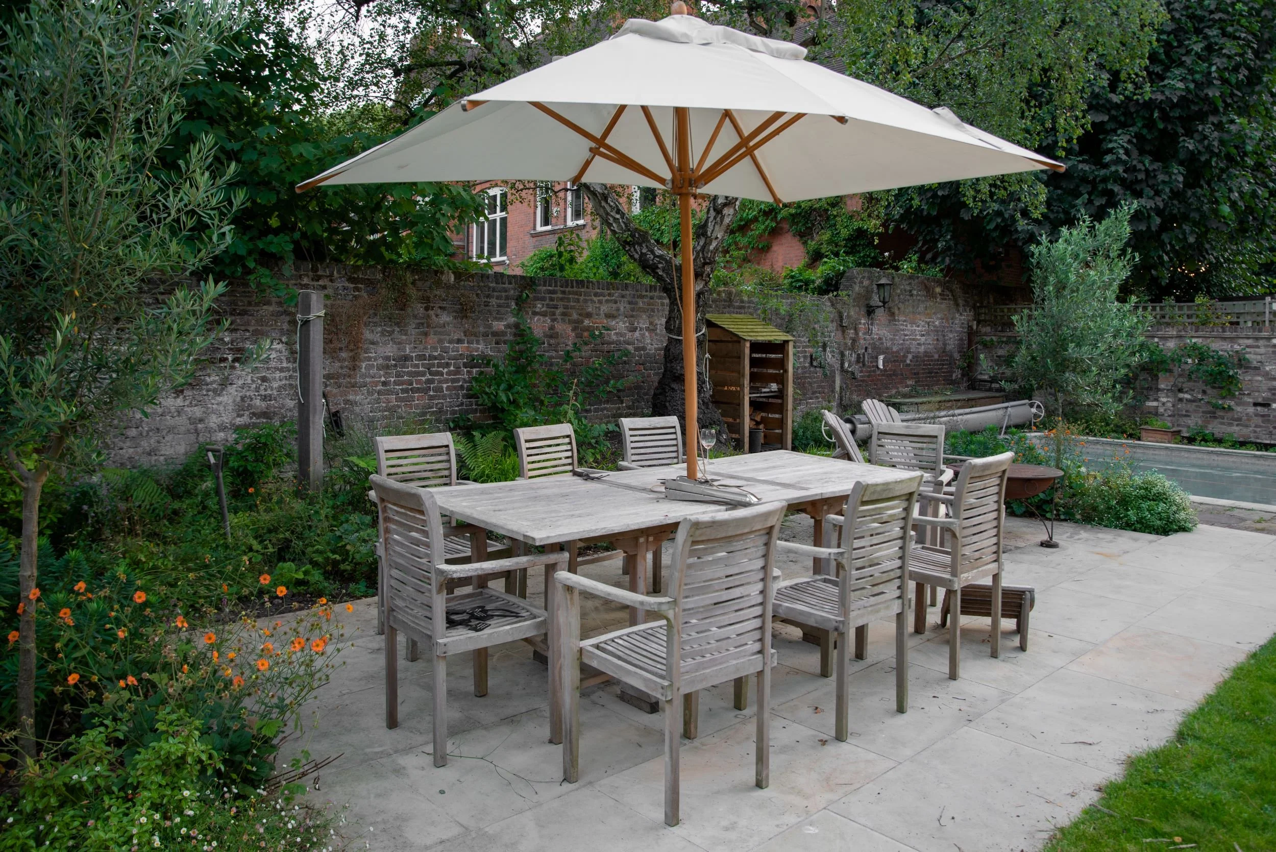 A teak dining table and chairs sit beneath a large parasol on a stone terrace in a Wimbledon Village garden. JaneShanksterGardenDesign.co.uk

A generous teak dining set for eight is shaded by a square canvas umbrella on a paved terrace, with olive tr