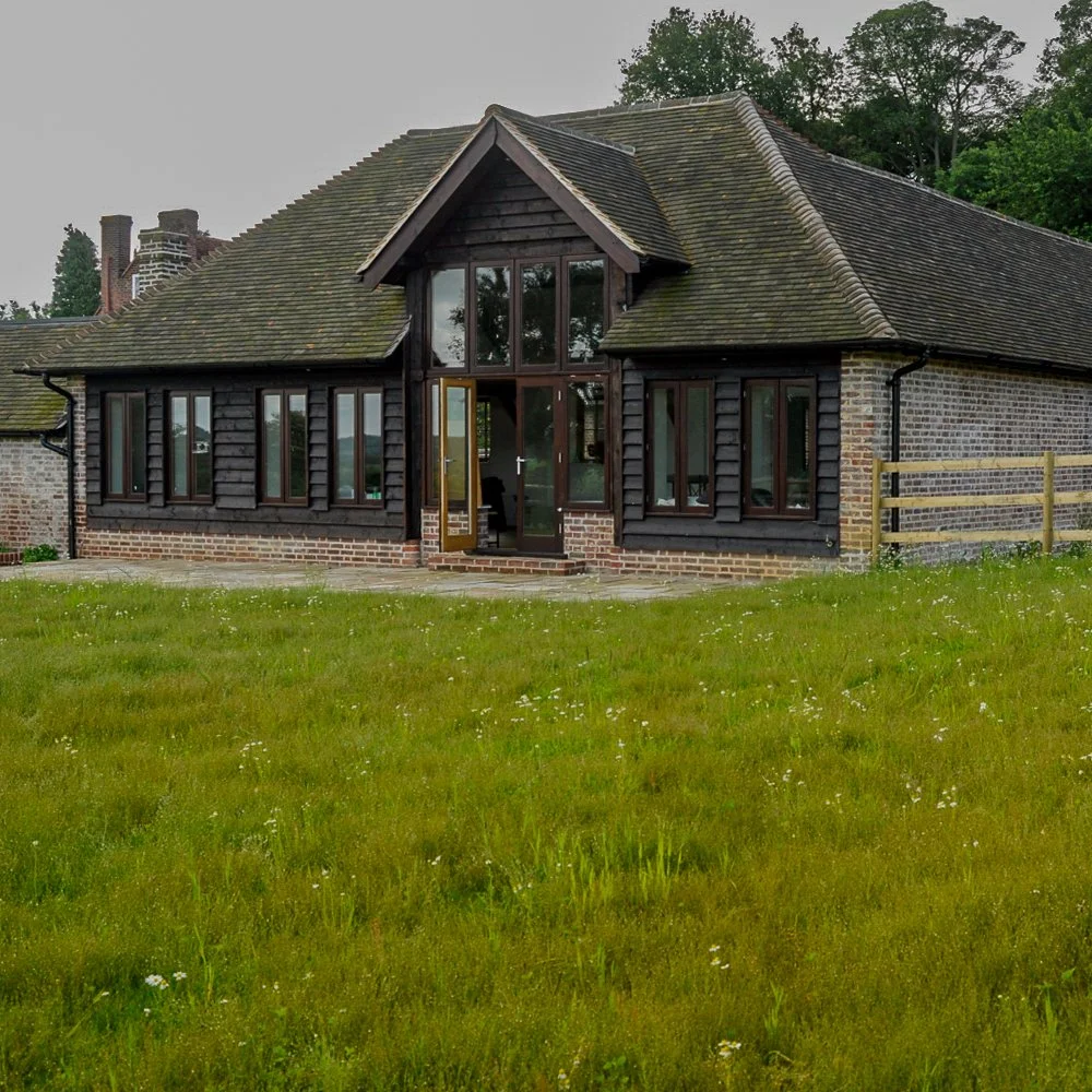 An oak-framed brick house in Surrey stands behind a plain, unplanted meadow-like lawn before its garden redesign. JaneShanksterGardenDesign.co.uk.

This is the garden before any planting took shape: a handsome oak-framed house, a former dairy buildin