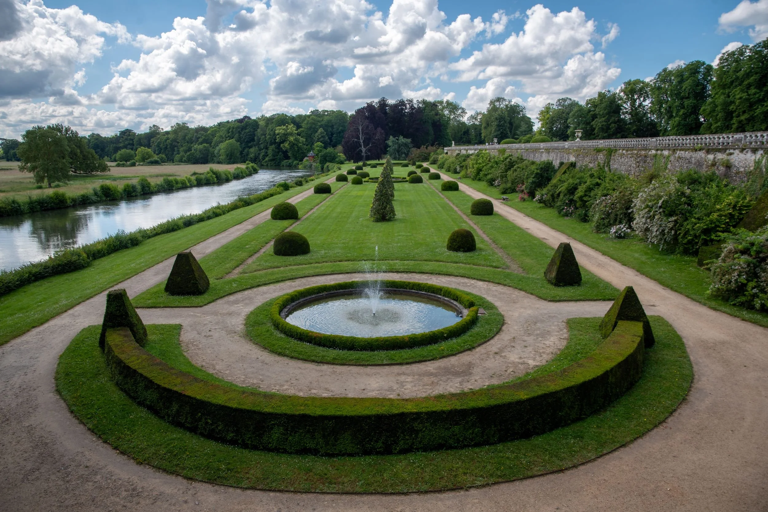 Formal lower garden at Château du Lude with a circular fountain, clipped box hedging and pyramid topiary, the River Loir running alongside.
