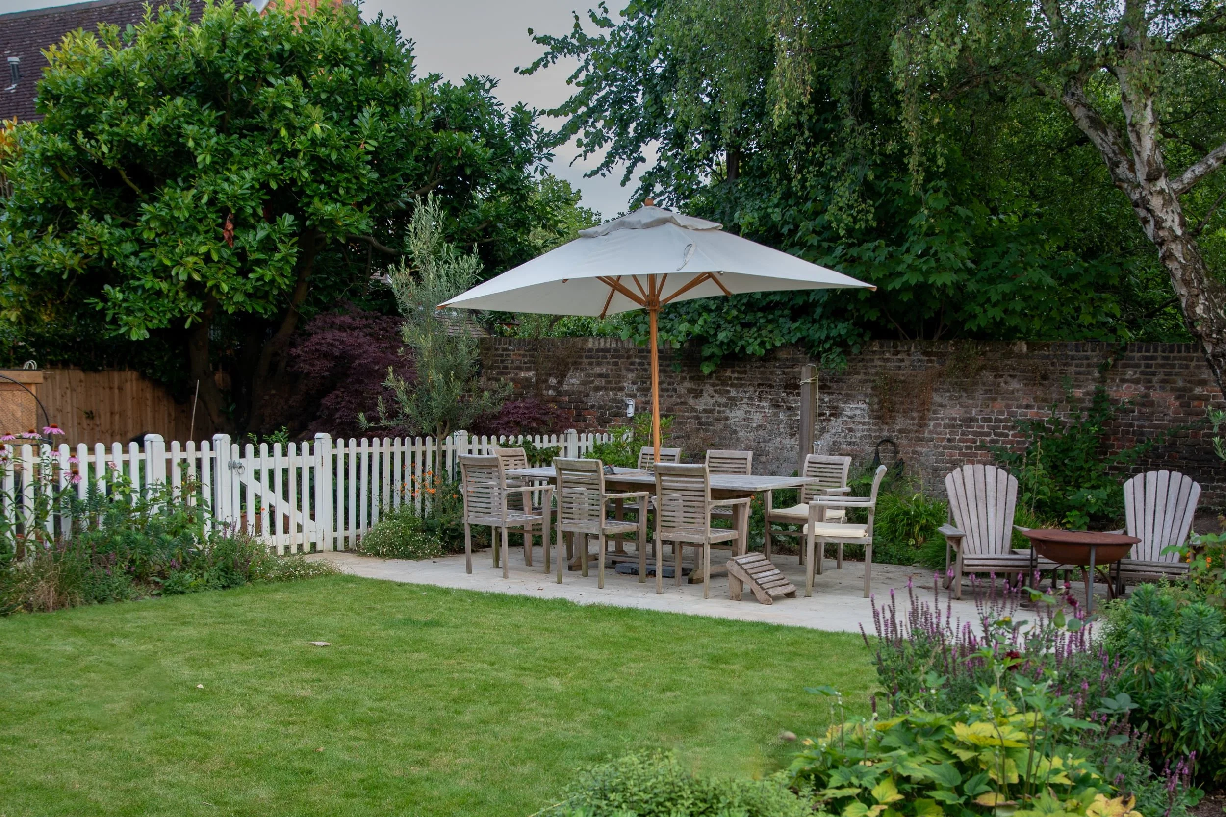 A wide view of a Wimbledon Village garden showing the dining terrace, firepit seating area and white picket fence. JaneShanksterGardenDesign.co.uk

A broad view across the lawn shows the full outdoor layout: a shaded dining table for eight, a separat