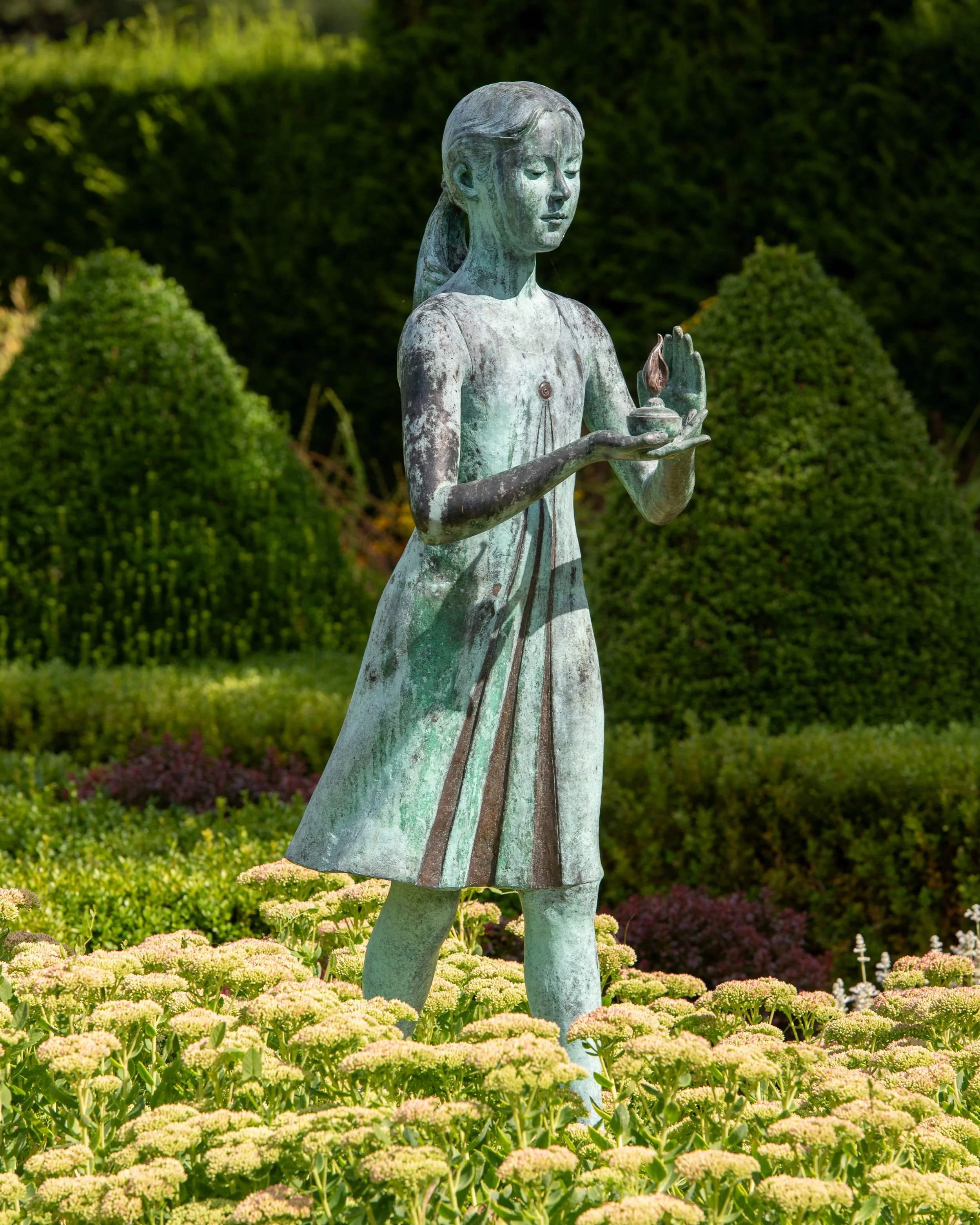 A bronze statue of a girl holding a lamp stands among sedum flowers, framed by clipped yew hedging at Waterperry Gardens