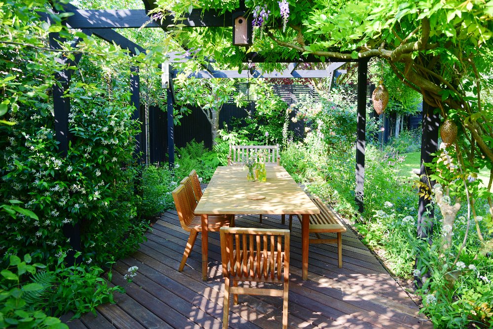 A wooden table and chairs sit in dappled sunlight beneath a pergola covered in star jasmine, wisteria and climbing roses in a Teddington garden. JaneShanksterGardenDesign.co.uk.

A wooden table and chairs sit in dappled sunlight beneath a pergola cov