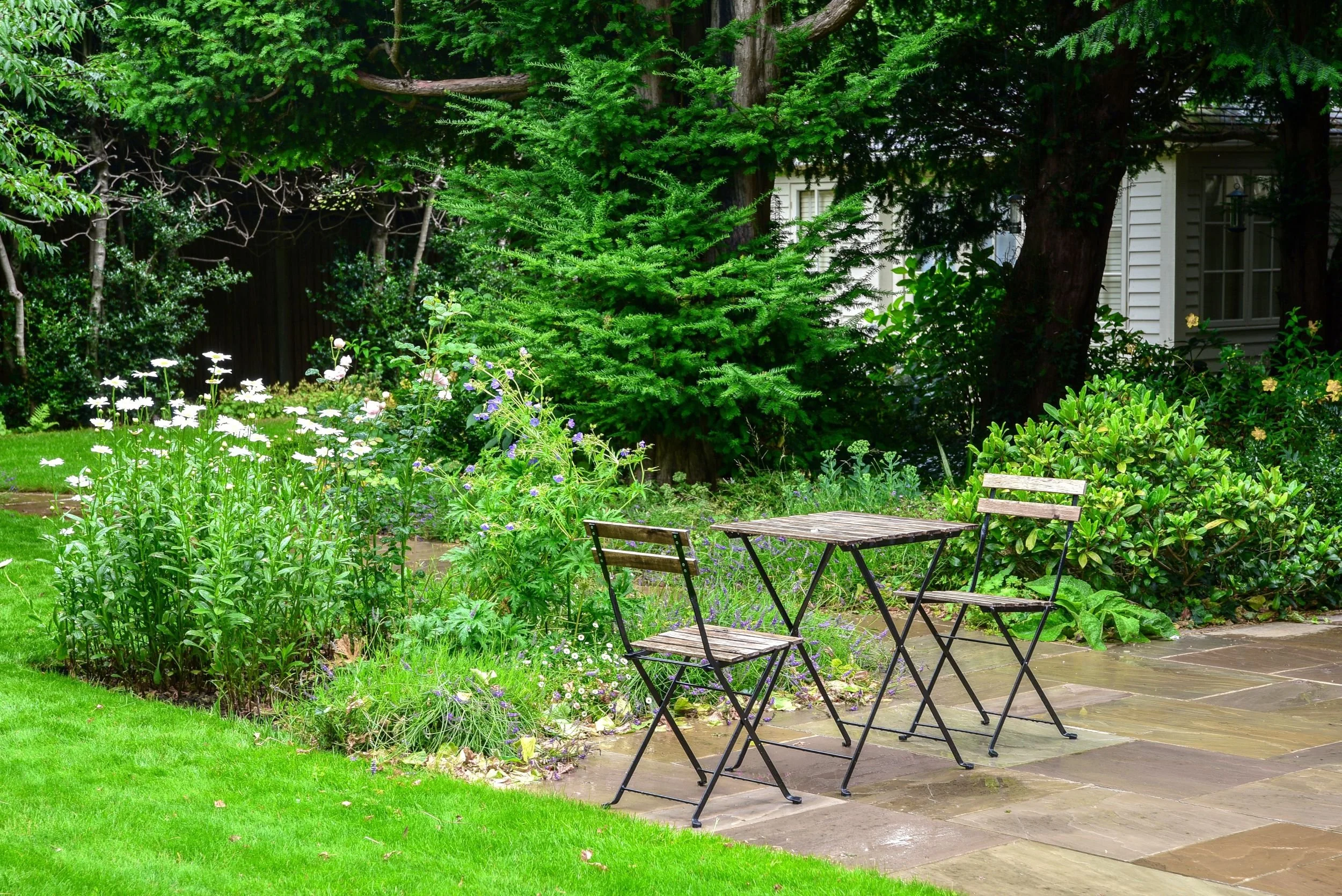 A small bistro table and chairs sit on a stone terrace beside a border of shasta daisies and roses. JaneShanksterGardenDesign.co.uk

A simple wrought-iron bistro set offers a quiet coffee spot on the stone terrace, tucked beside a border of white sha