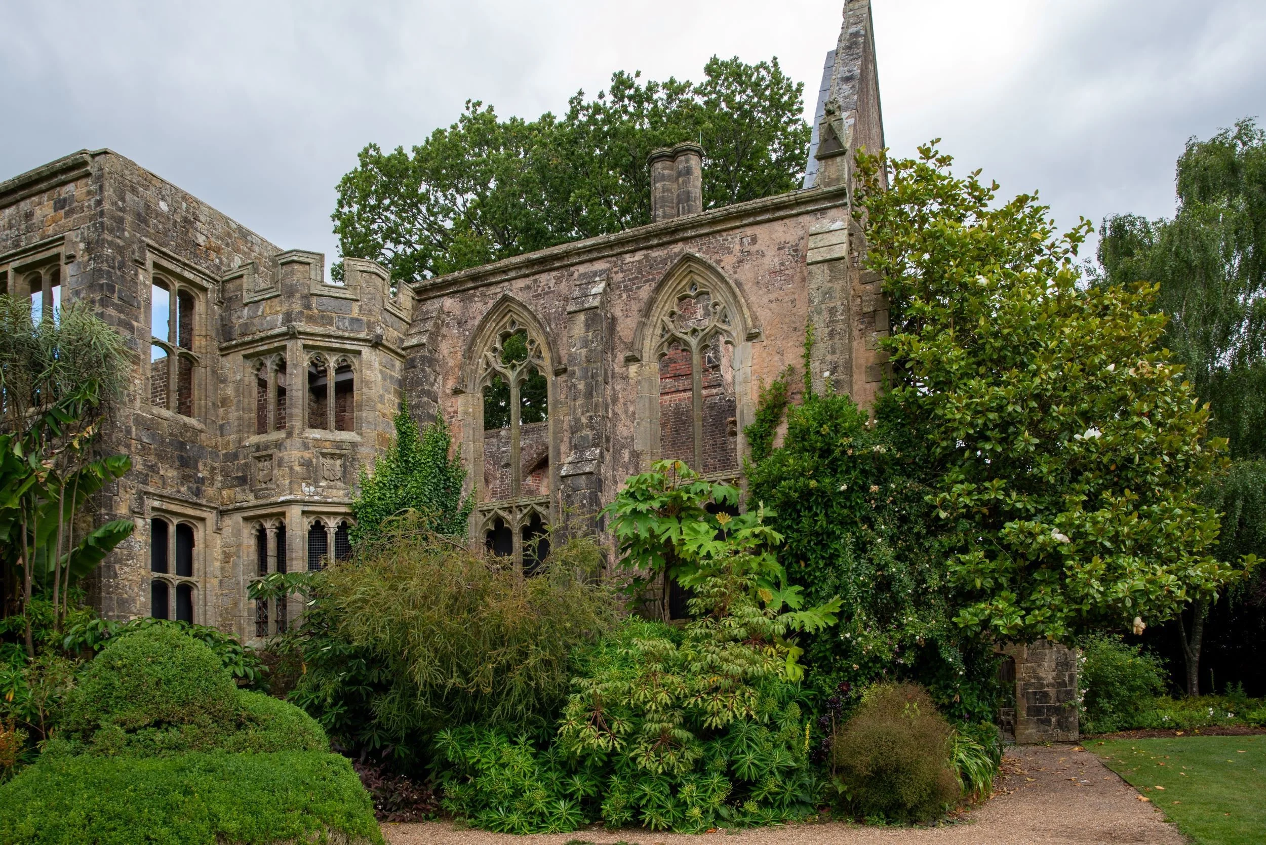 Ivy-clad ruins of Nymans house showing Gothic tracery windows and a castellated tower, surrounded by mature planting.