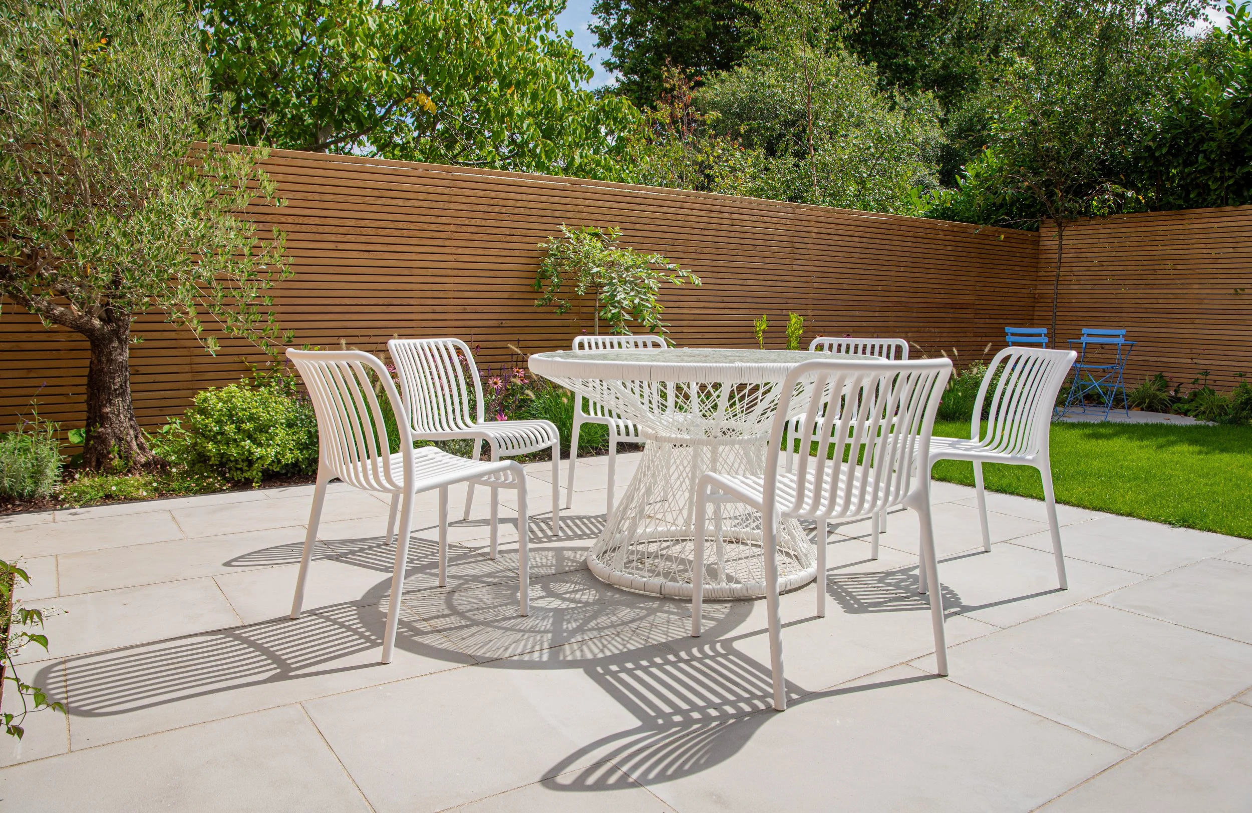 White dining chairs beside olive tree and echinacea border in property development garden

A close view of white woven-style dining chairs set against a backdrop of cedar slat fencing, a mature olive tree, and a vibrant border of echinacea, salvia an