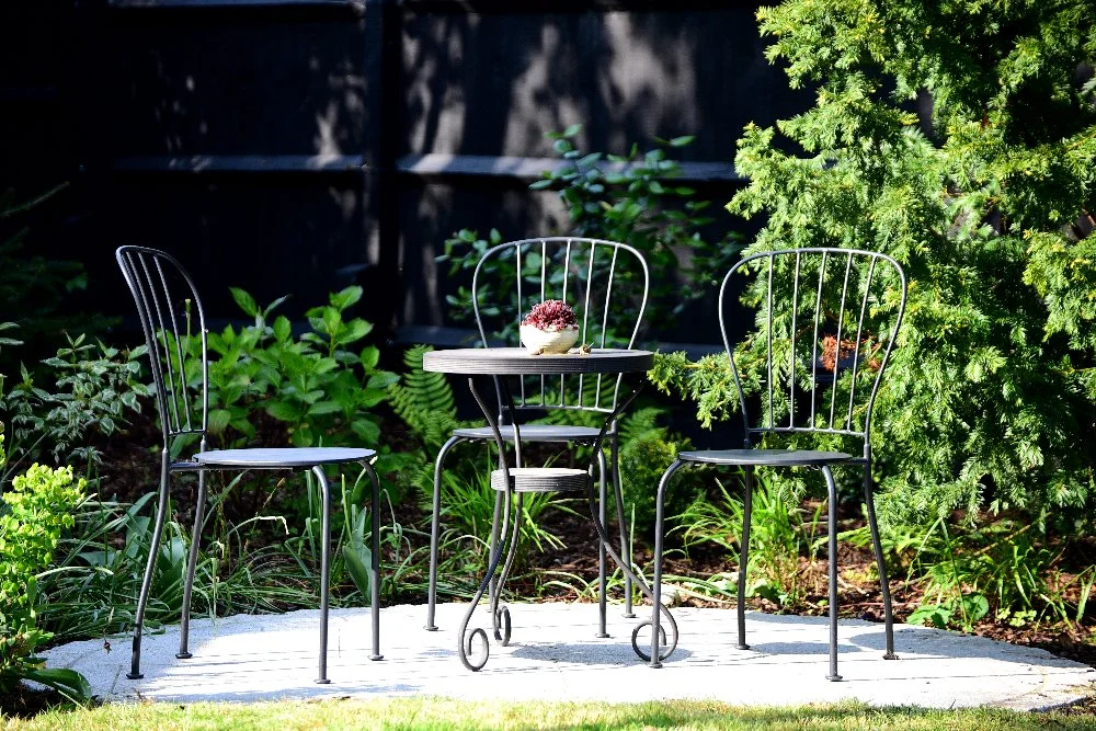Metal bistro table and chairs on garden patio, Wimbledon garden by Jane Shankster Garden Design

A slender metal bistro table and chairs occupy a sunlit paved corner, tucked beside a conifer and lush green planting. This intimate seating spot offers 