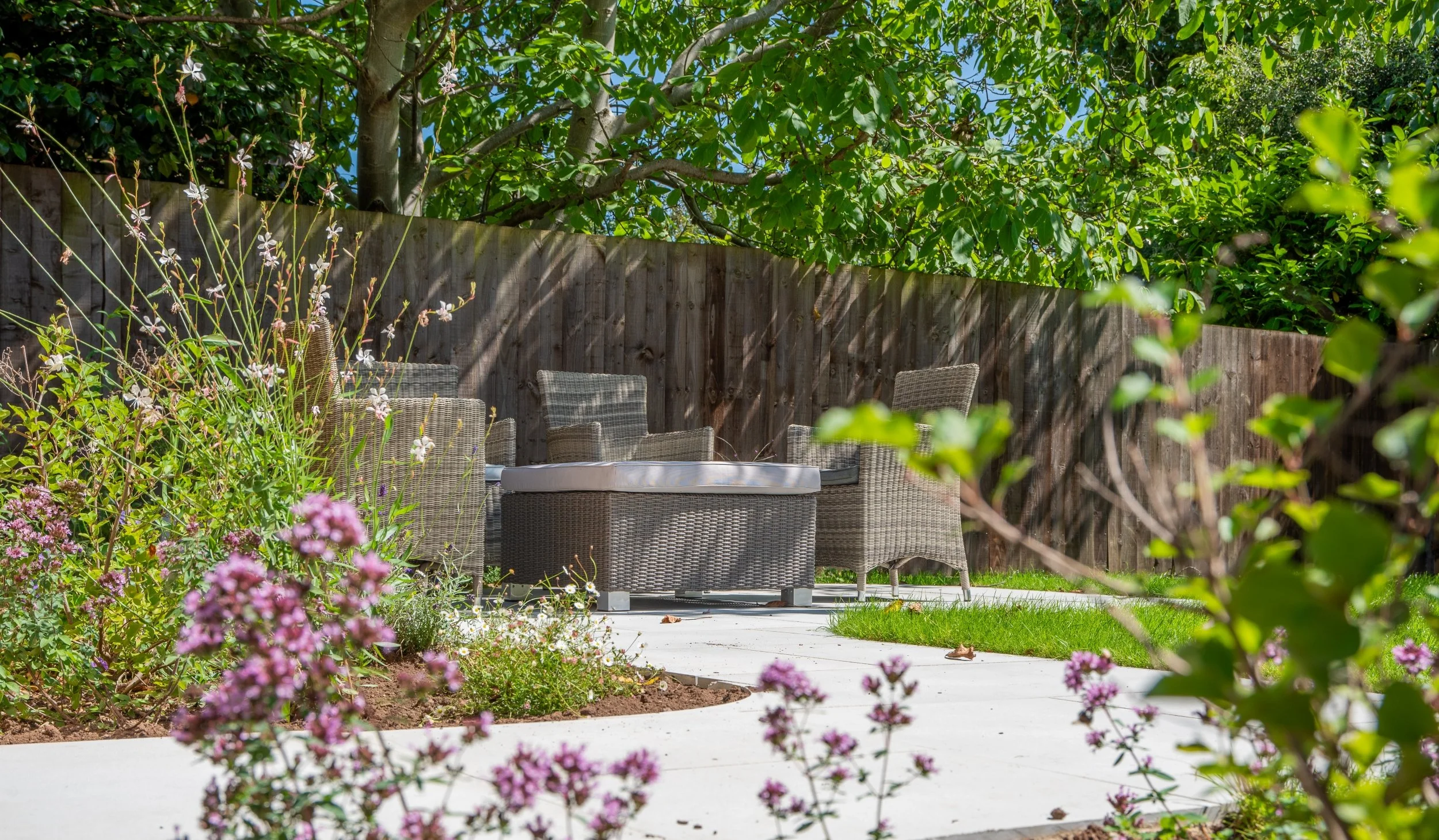 Rattan armchairs and ottoman glimpsed through flowering border in property development garden

A woven rattan seating set with a cushioned ottoman, framed by a soft curtain of gaura and oregano in full flower. A curved stone path leads through the pl