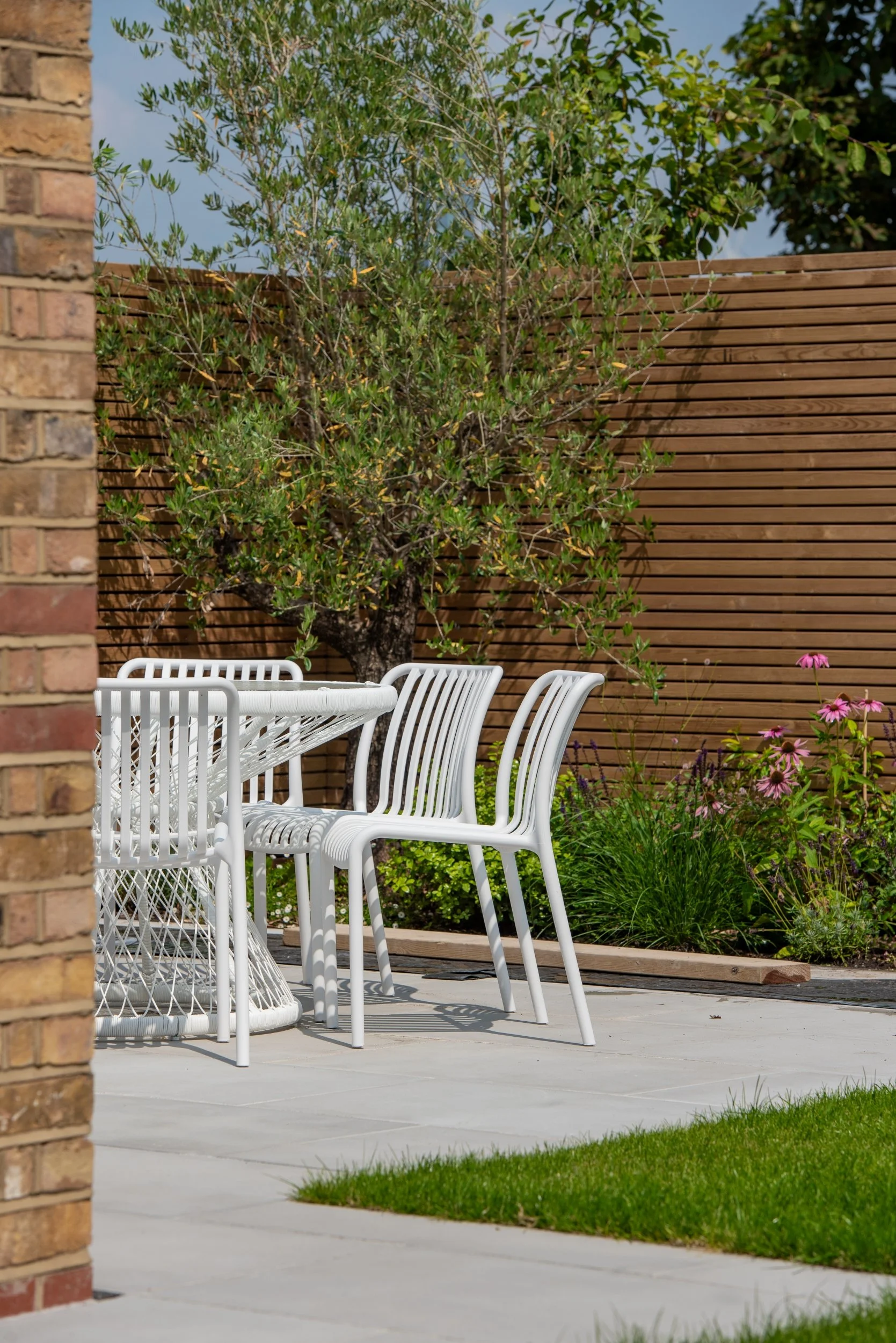 White dining chairs beside olive tree and echinacea border in property development garden

A close view of white woven-style dining chairs set against a backdrop of cedar slat fencing, a mature olive tree, and a vibrant border of echinacea, salvia an