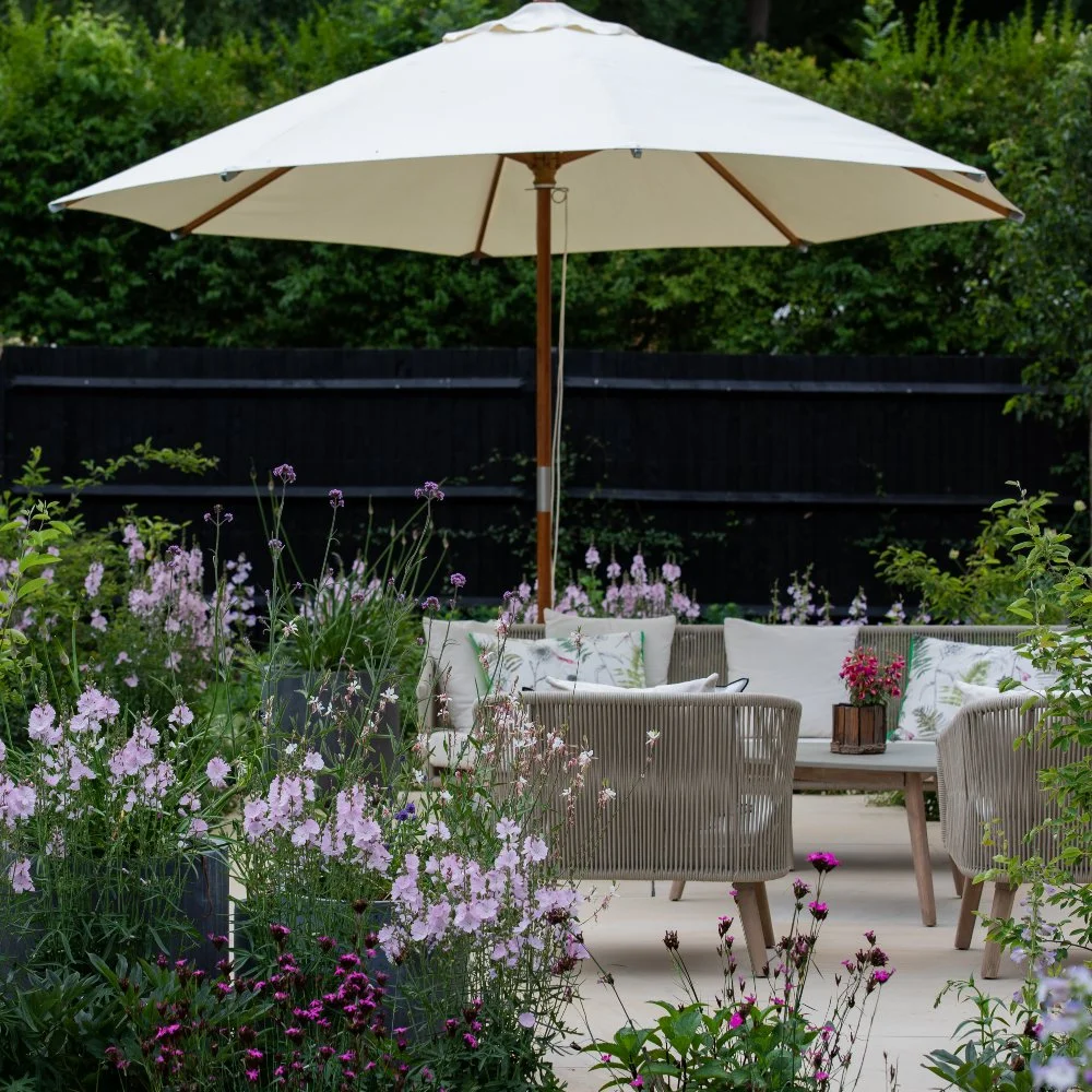 A stone terrace with rope-weave garden furniture and a large cream parasol in a Surrey country garden, framed by abundant borders of pink sidalcea, purple verbena and white gaura against a black timber fence. ArthurRoadLandscapes.co.uk. 

A second vi