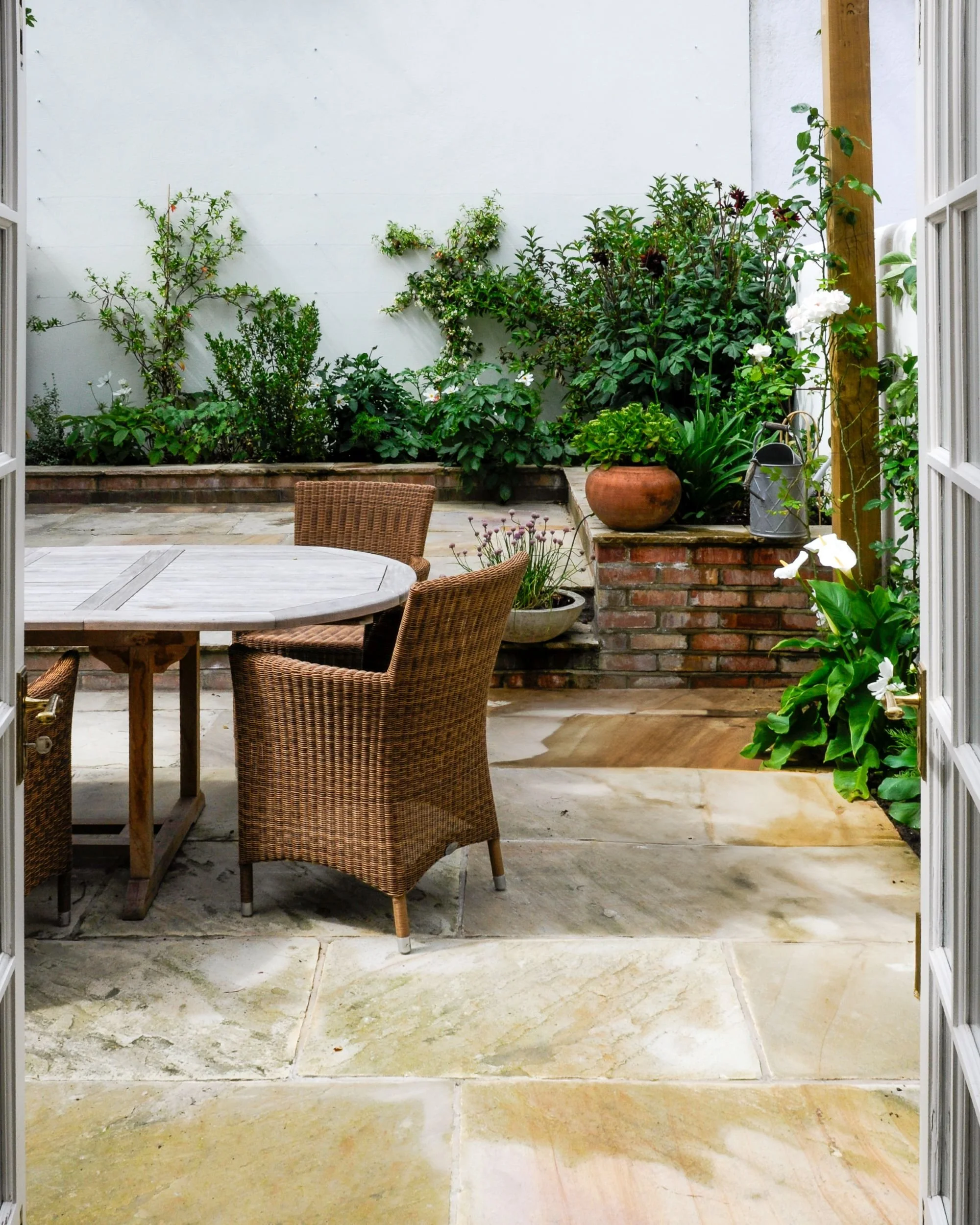View through open French doors onto a paved courtyard garden with a round teak table and wicker chairs

Looking out from the house through open French doors, sandstone paving leads to a round teak table and wicker armchairs, framed by raised brick bo