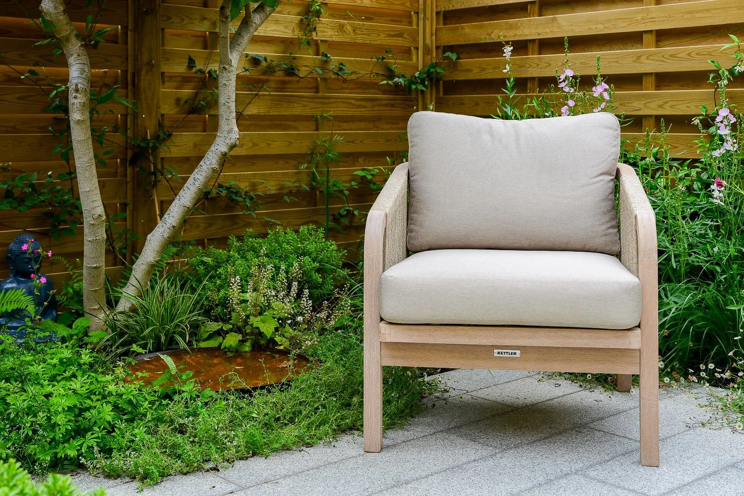Close-up of a lounge chair beside a Buddha statue and multi-stem tree in a courtyard garden

A close view of the seating corner, where a Kettler lounge chair sits beside a small Buddha statue beneath a multi-stem tree, framed by timber screening and 