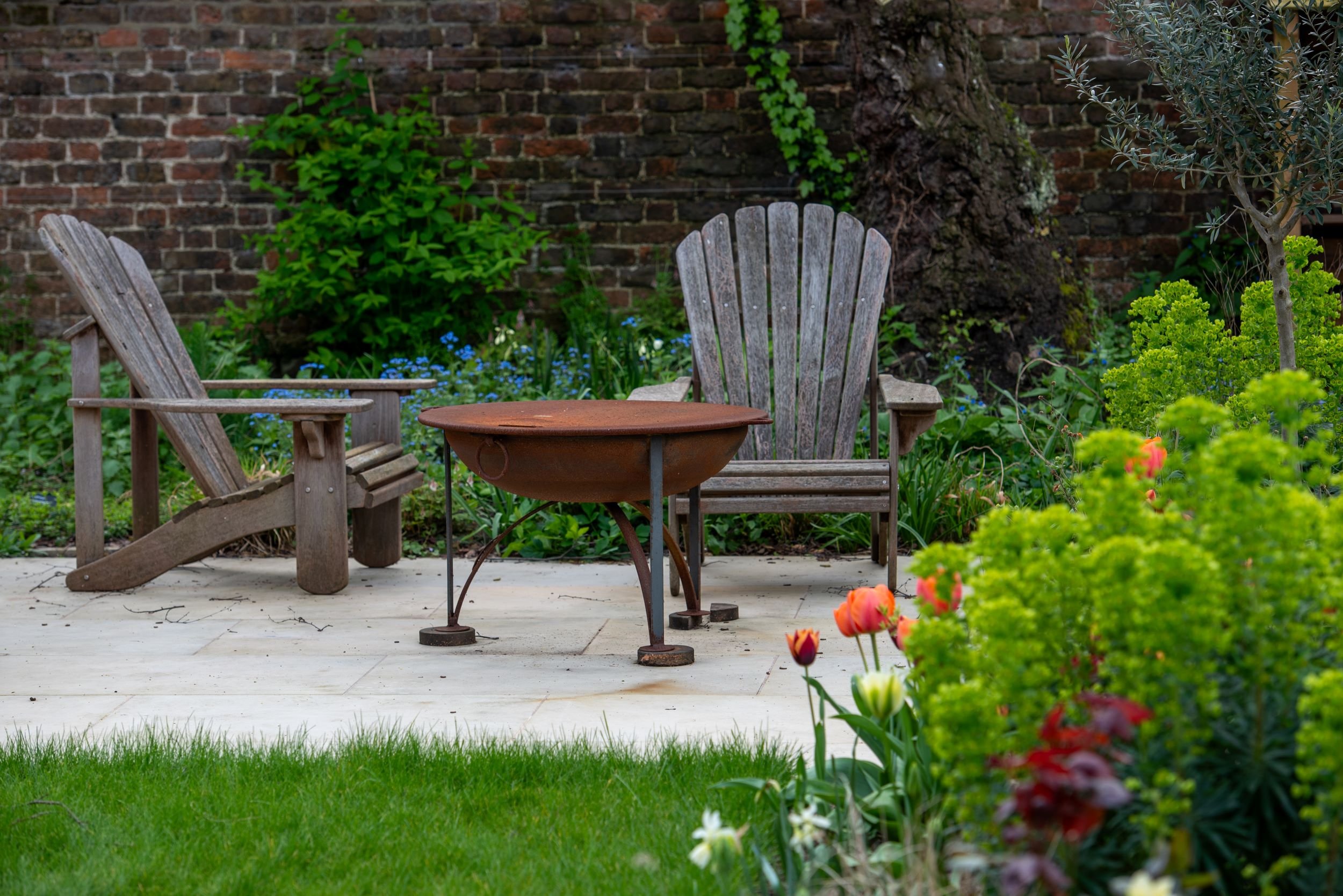 A rusted steel firepit sits between two weathered Adirondack chairs on a terrace surrounded by tulips and euphorbia. JaneShanksterGardenDesign.co.uk

A pair of grey-weathered Adirondack chairs flank a round corten steel firepit on stone paving, with 