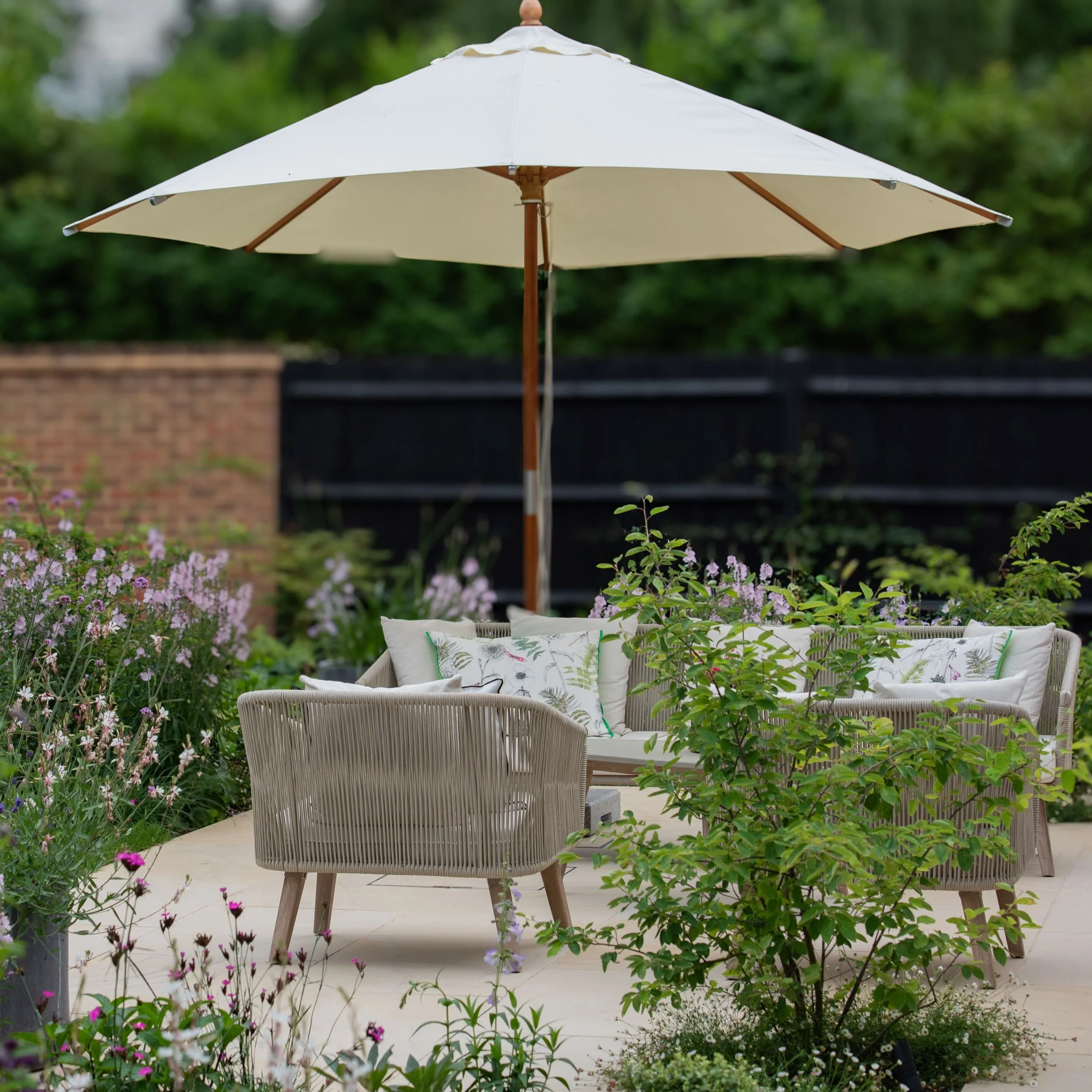A stone terrace seating area with rope-weave chairs, botanical print cushions and a large cream parasol, surrounded by naturalistic planting including gaura and sidalcea in a Surrey country garden. ArthurRoadLandscapes.co.uk.

 The main terrace seati