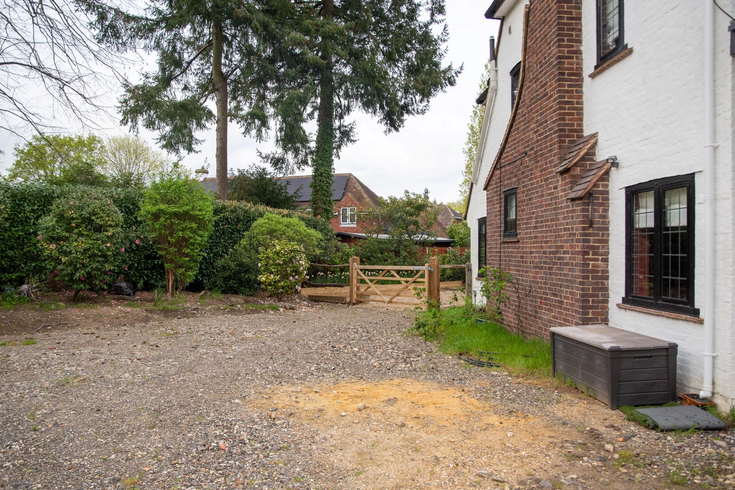 A large empty gravel driveway beside a brick and white house, with a timber five-bar gate and mature shrubs beyond.